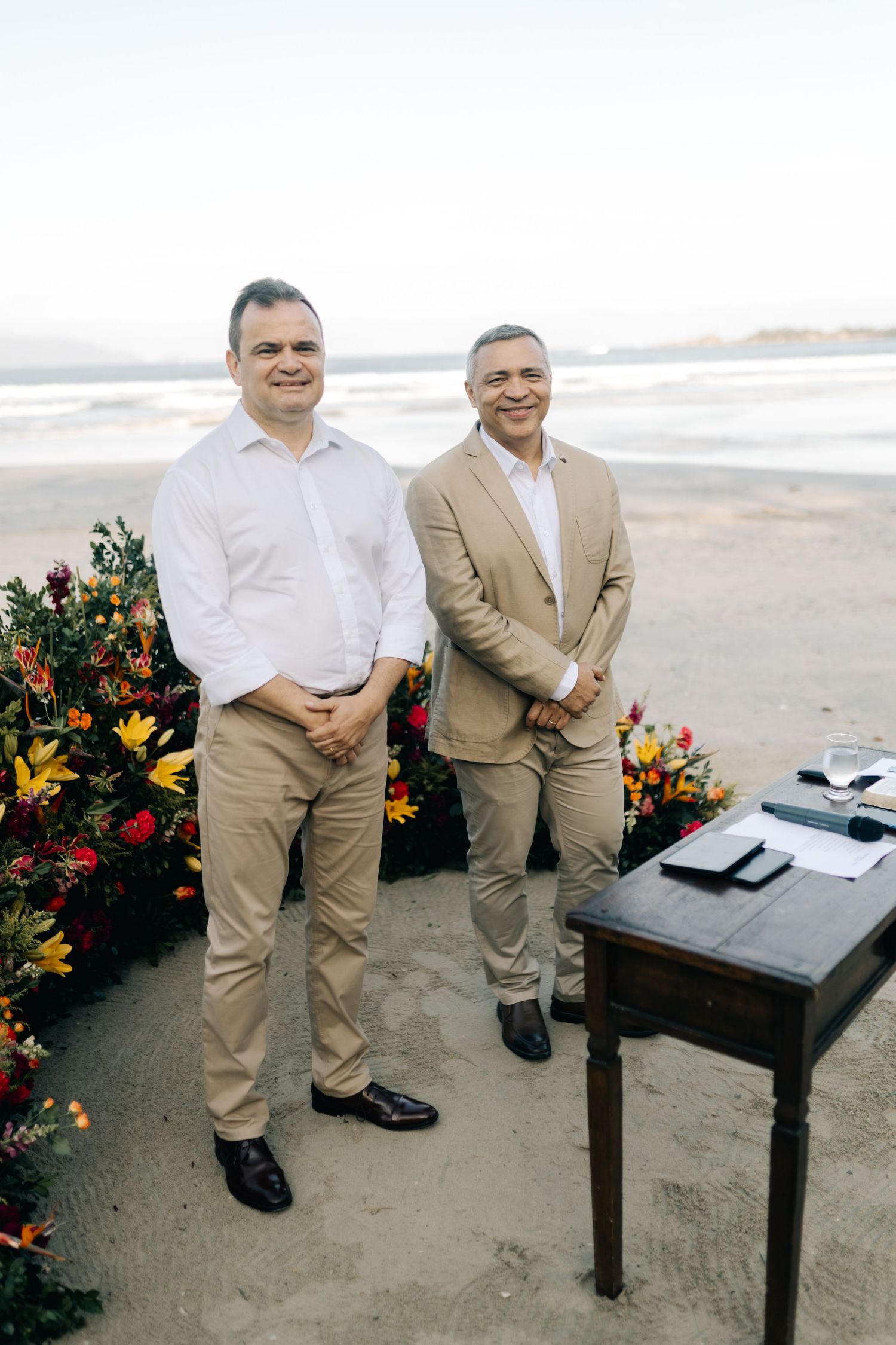 Two people in beige suits sitting at a table on a sandy beach with floral decorations.