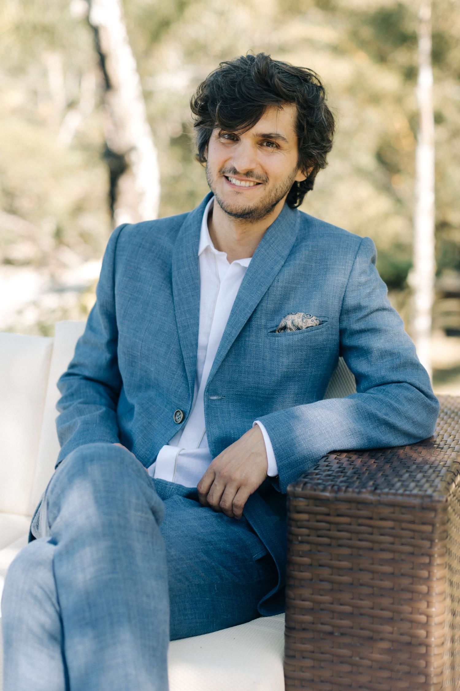 A portrait of someone in a blue suit seated on outdoor wicker furniture.