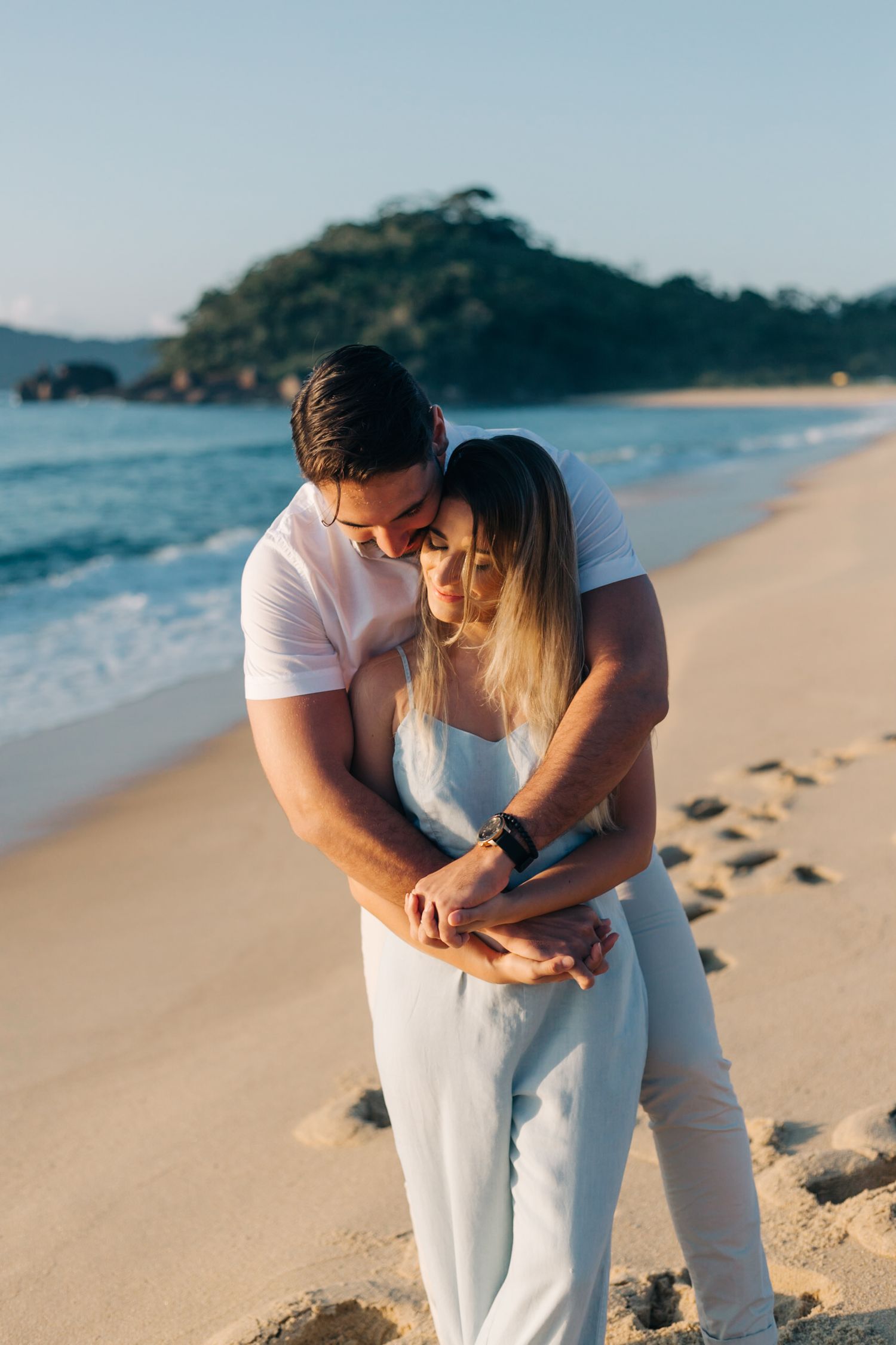 Intimate beachside moment between couple in white clothing with ocean waves in background.