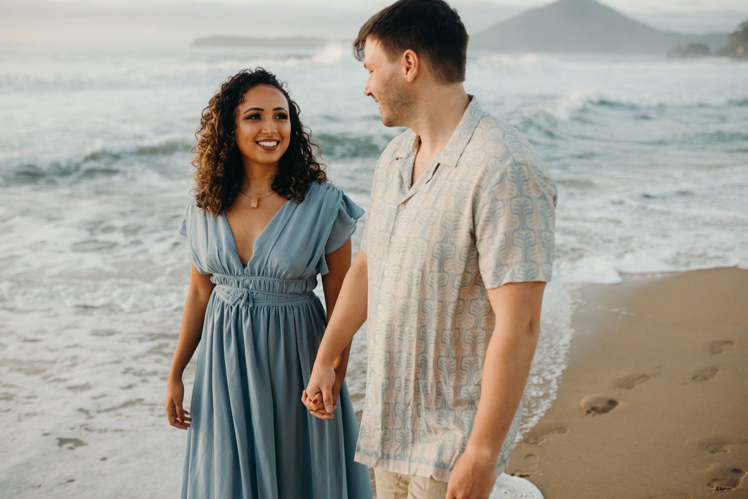 Happy couple laughing together on scenic beach with ocean waves and mountains.