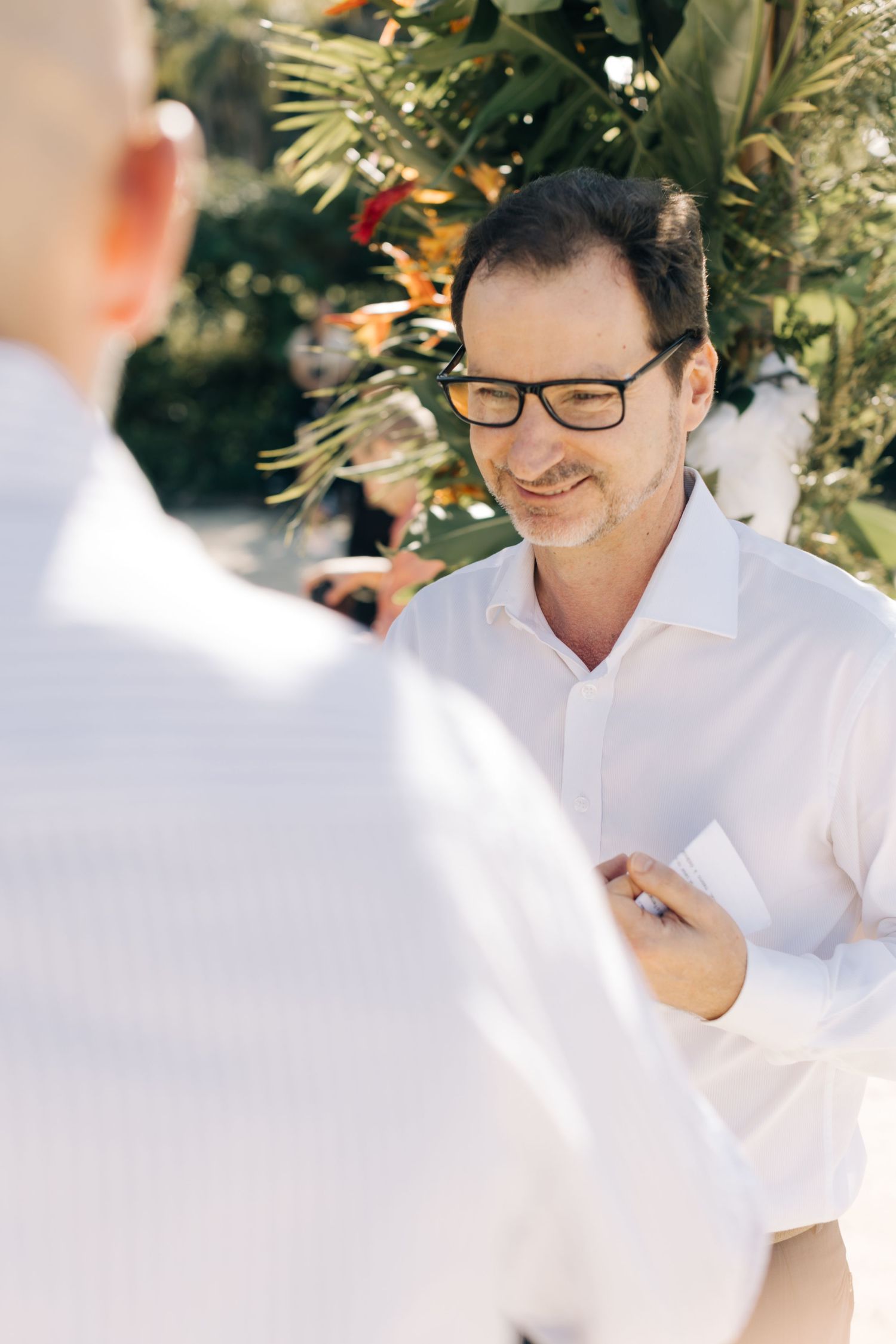 Person in white shirt and glasses smiling during outdoor wedding ceremony.