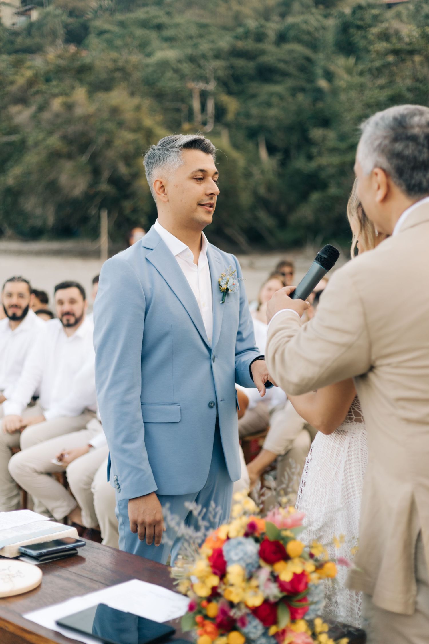 Outdoor wedding ceremony with guests in white attire and light blue suited groom at rustic table.