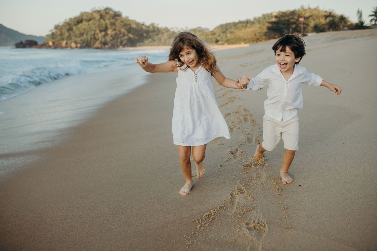 Kids in white outfits playing and running along the shoreline at sunset.