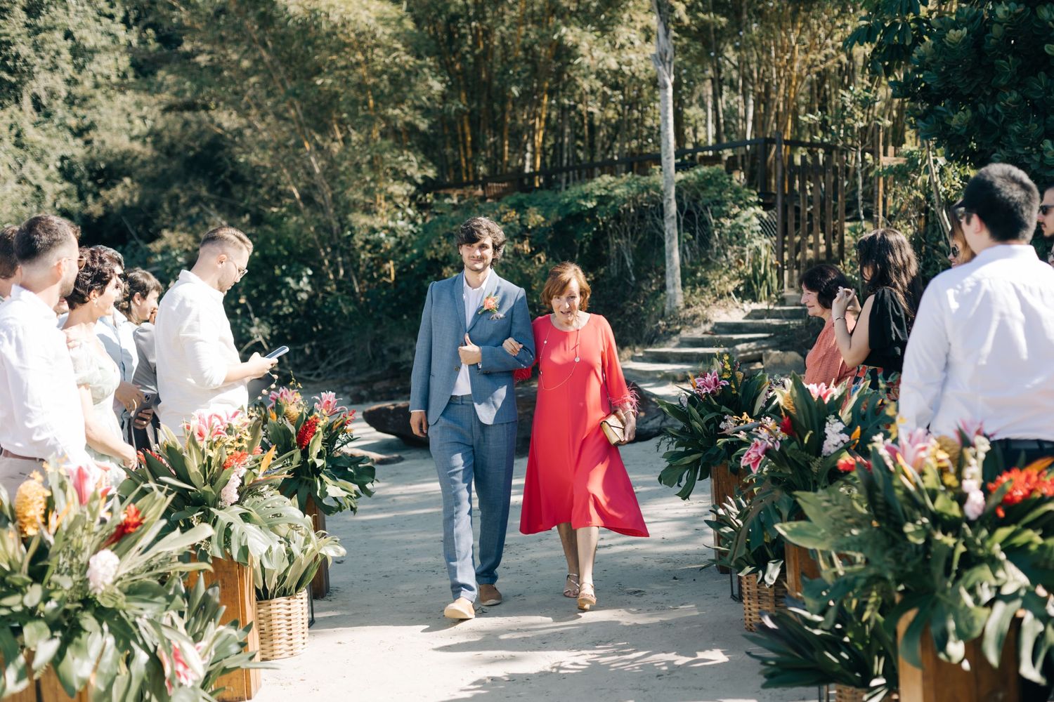 Outdoor wedding reception with guests mingling among tropical floral arrangements and greenery.