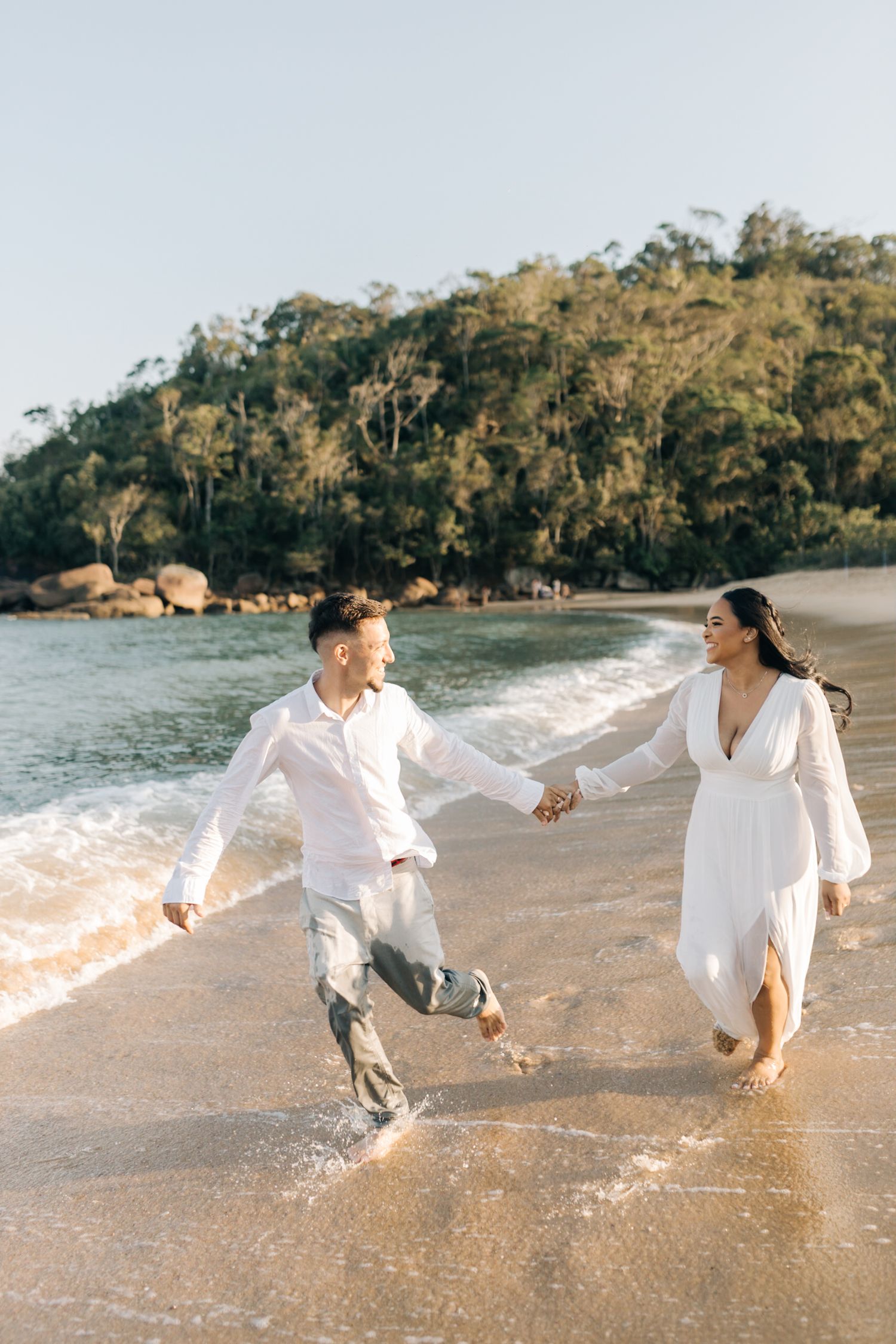 Energetic couple jumps and plays in beach waves while wearing matching white outfits.