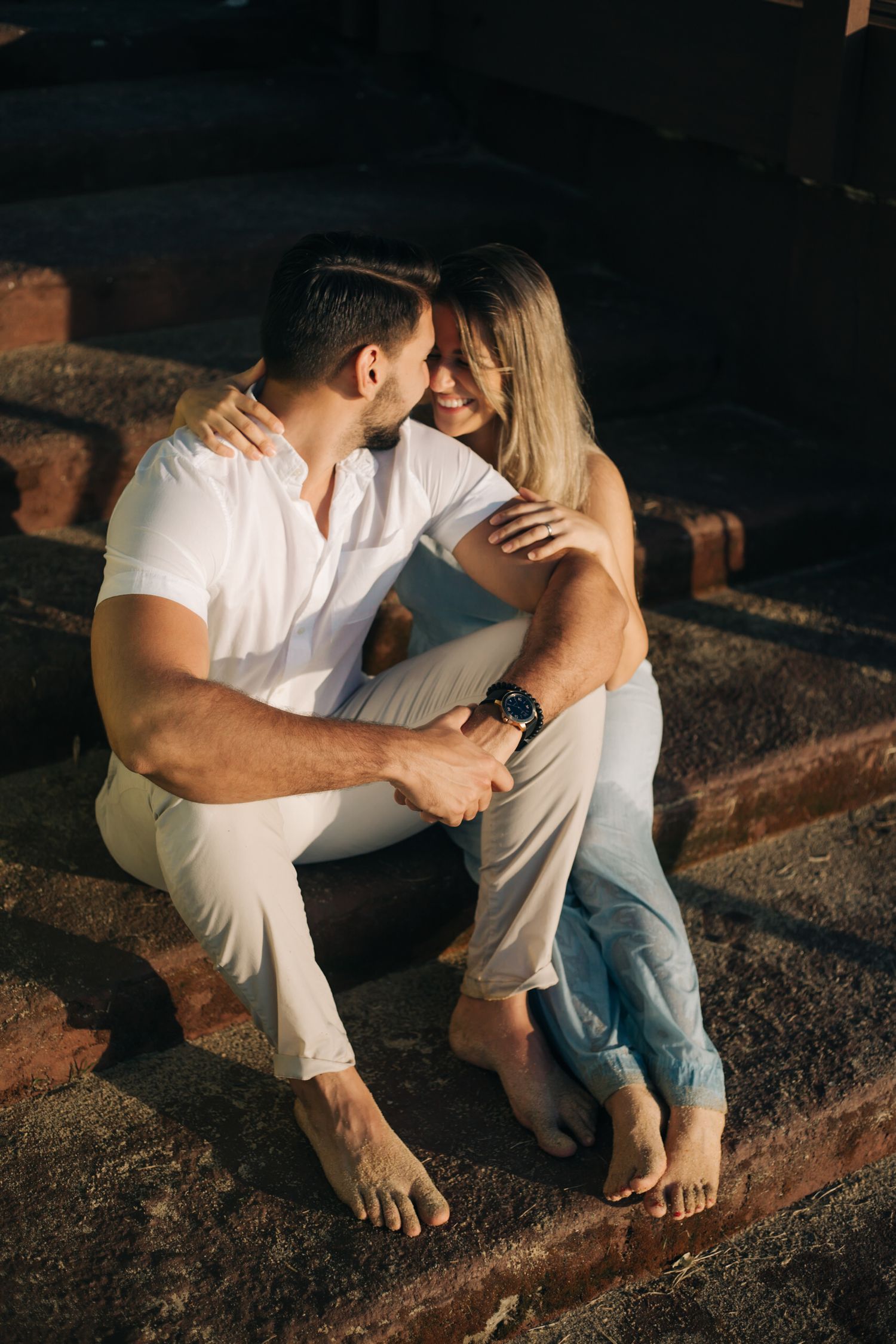 Tender moment between couple sitting on stone steps in casual white and denim outfits.