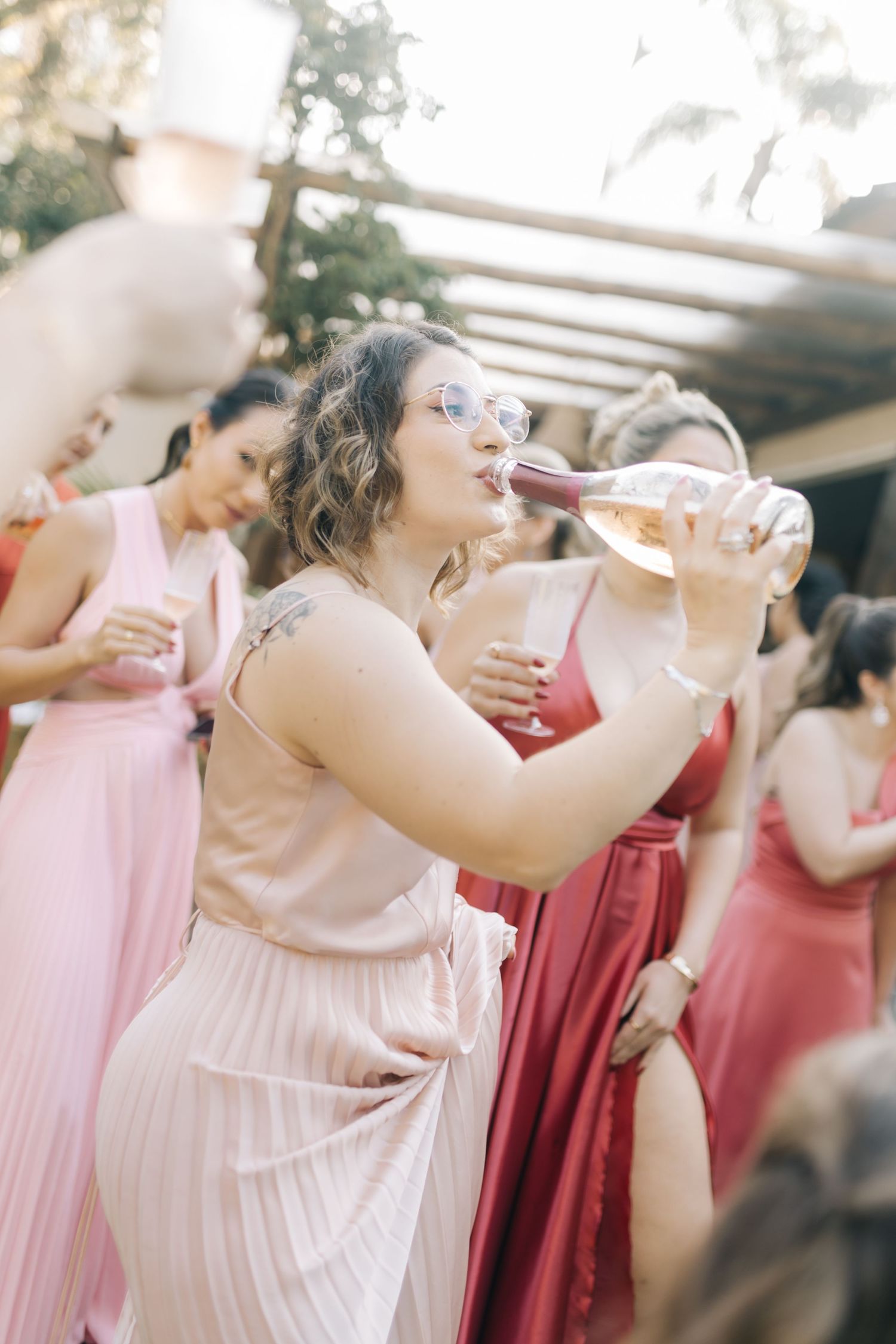Wedding guest in a pink dress enthusiastically drinks from a champagne glass during outdoor celebration.