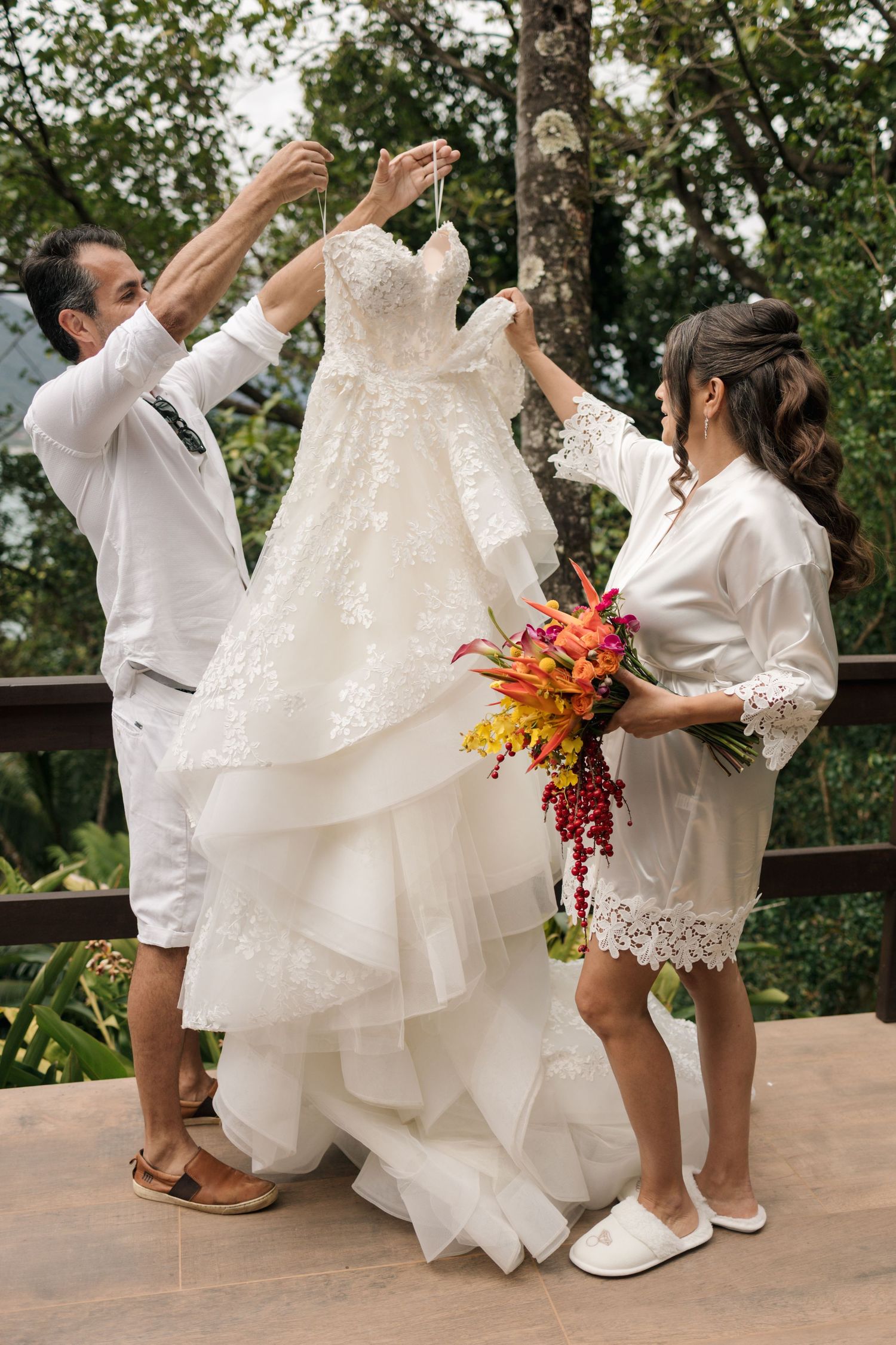 Wedding party in white robes helps fluff out layers of a bride's dress on an outdoor deck.