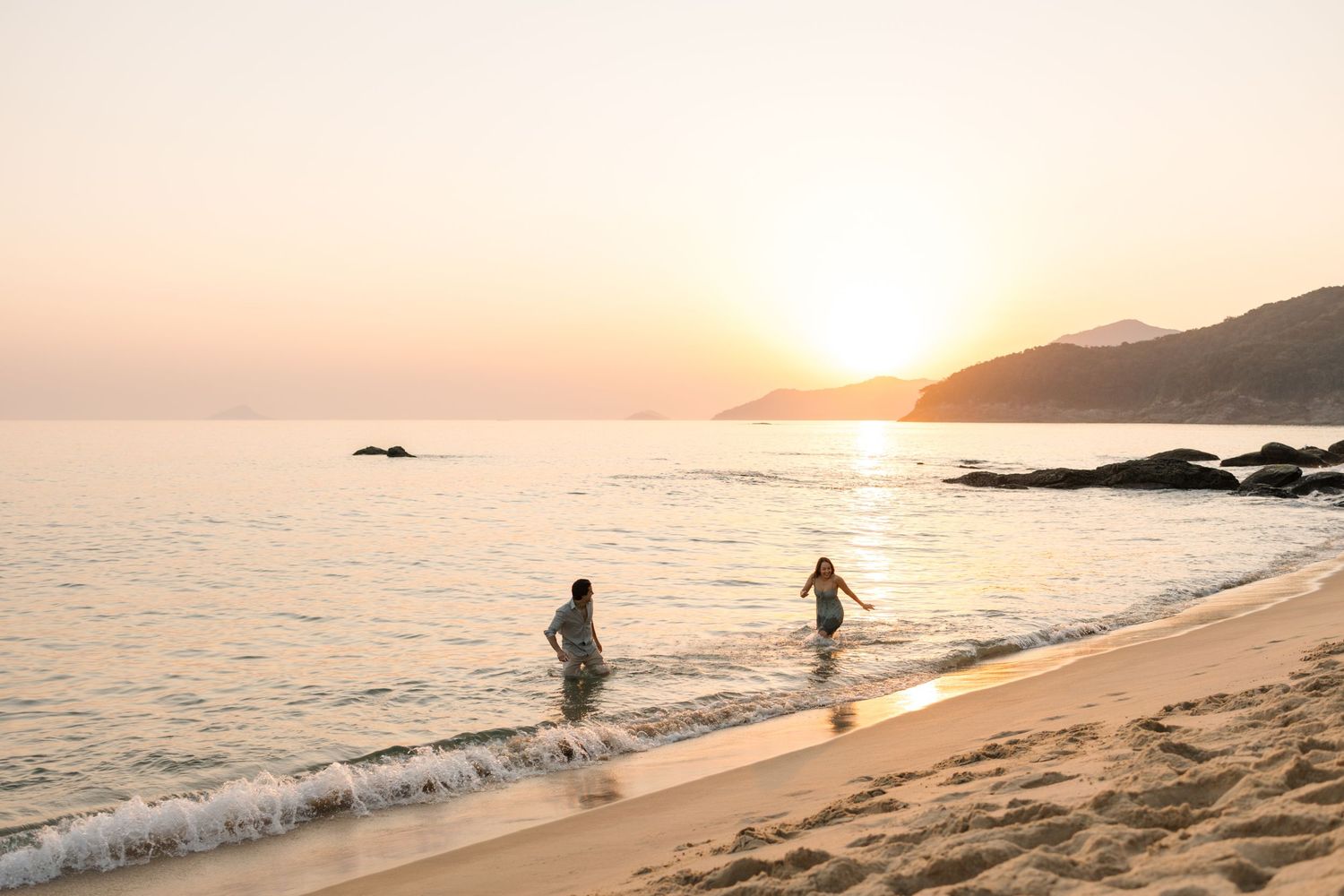 Scenic beach sunset with silhouettes of people walking along the shoreline and mountains in the distance.