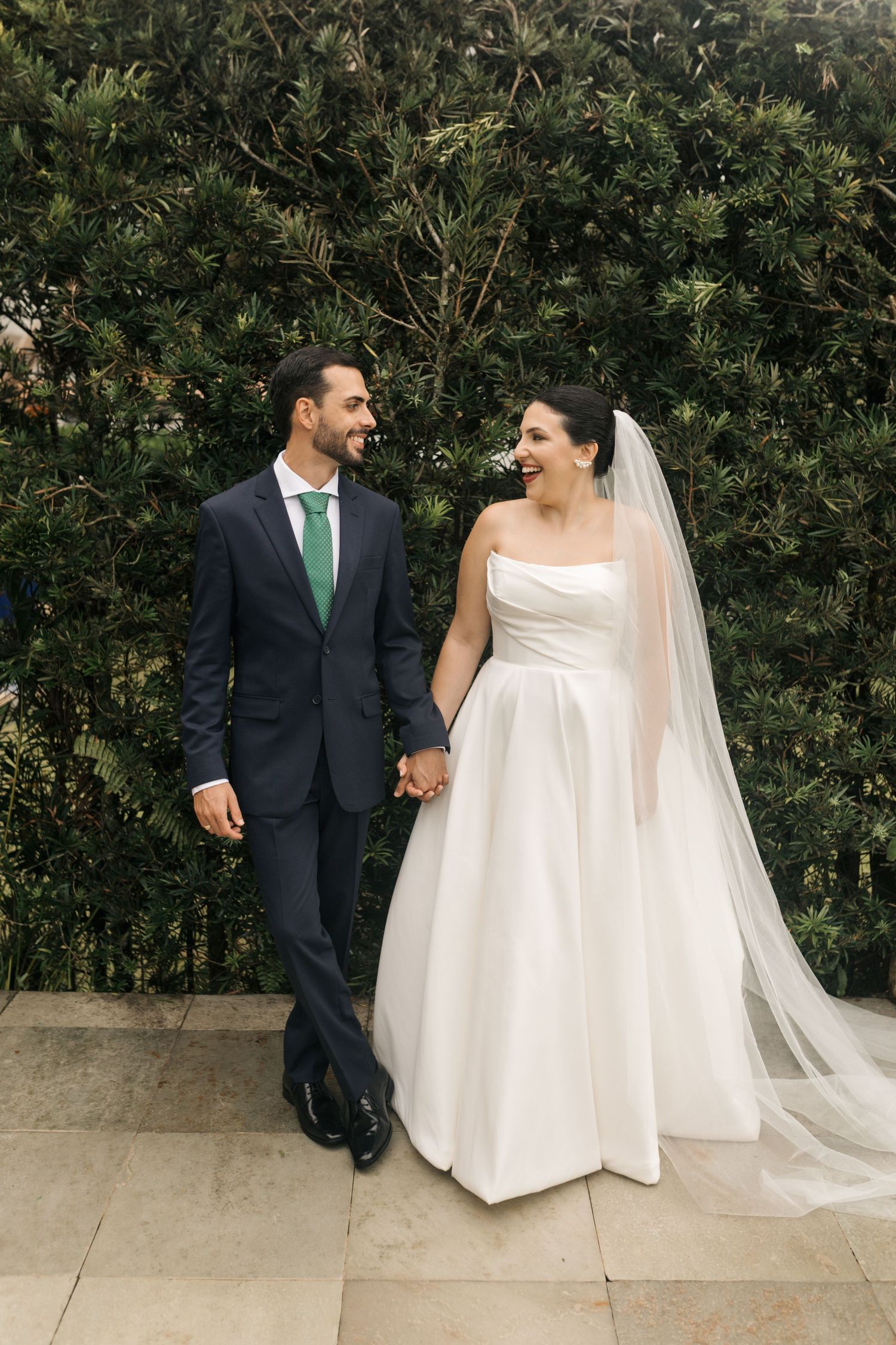 A wedding couple stands together against greenery, with the bride wearing a classic white gown and groom in navy suit.