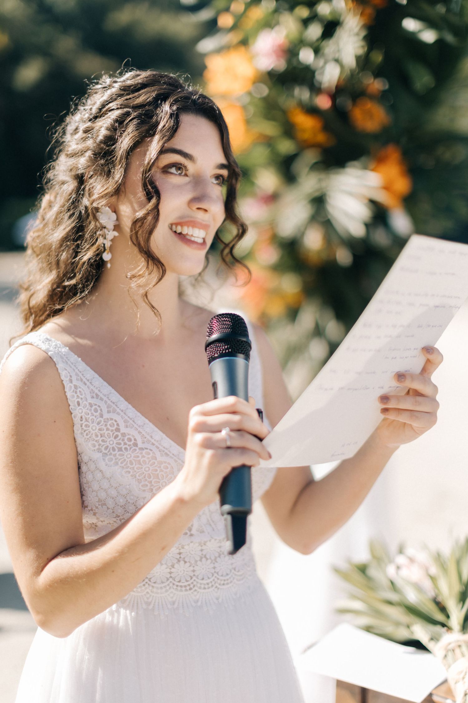 A bride in a lace wedding gown smiles while reading from paper during a beach ceremony.