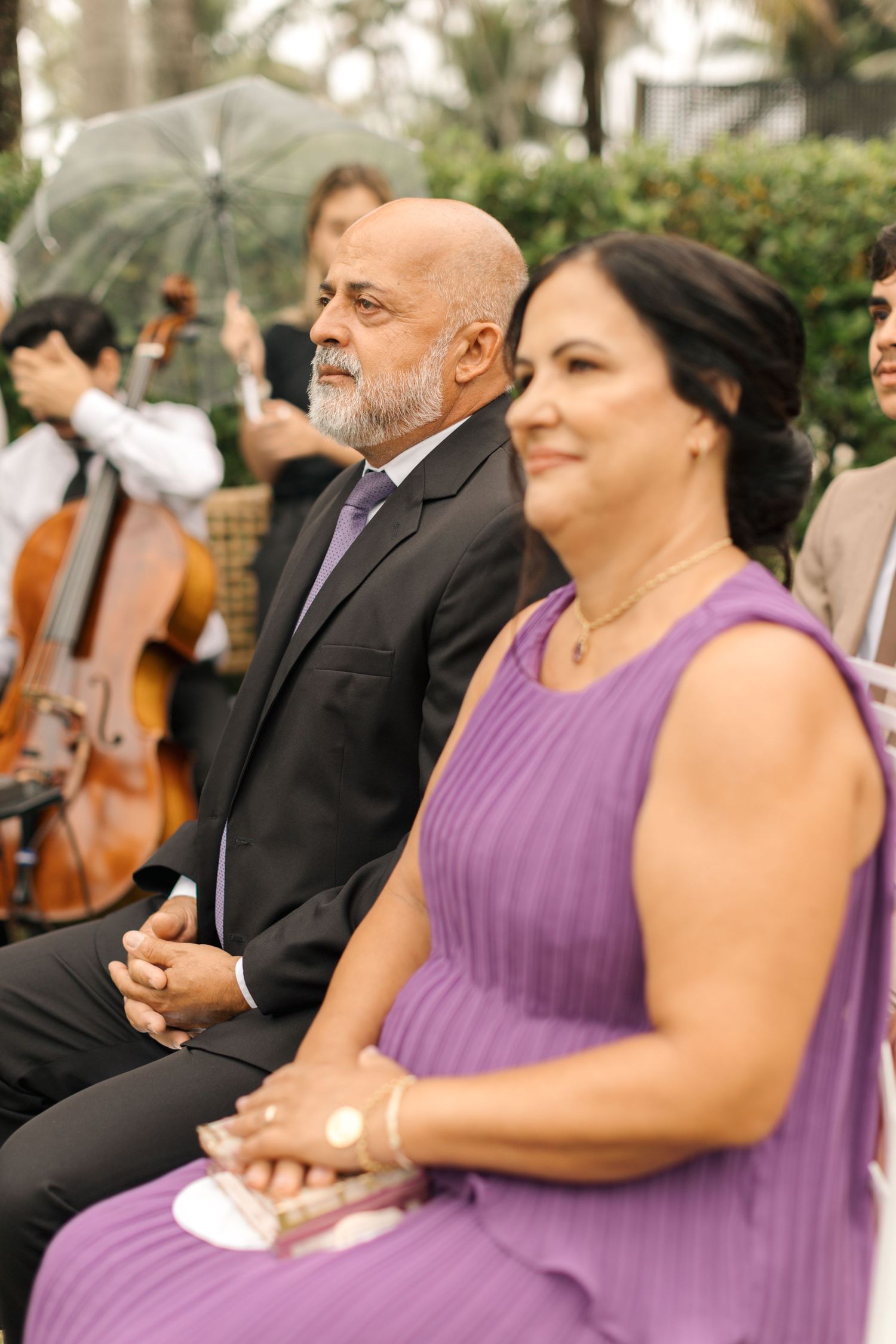 Two wedding guests in formal attire sit near a cellist during an outdoor ceremony.