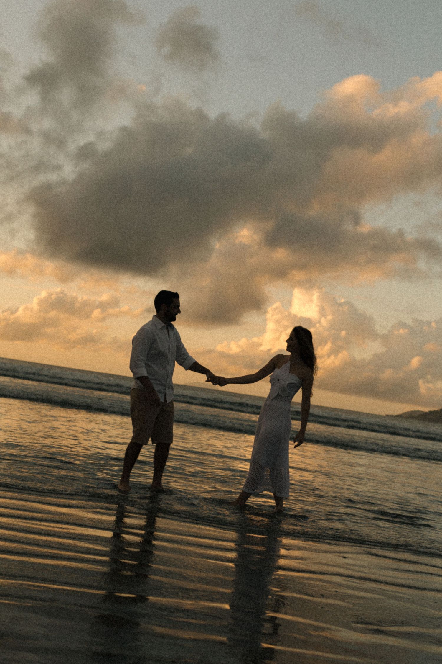 Two silhouettes hold hands while standing in shallow ocean water during a golden sunset.