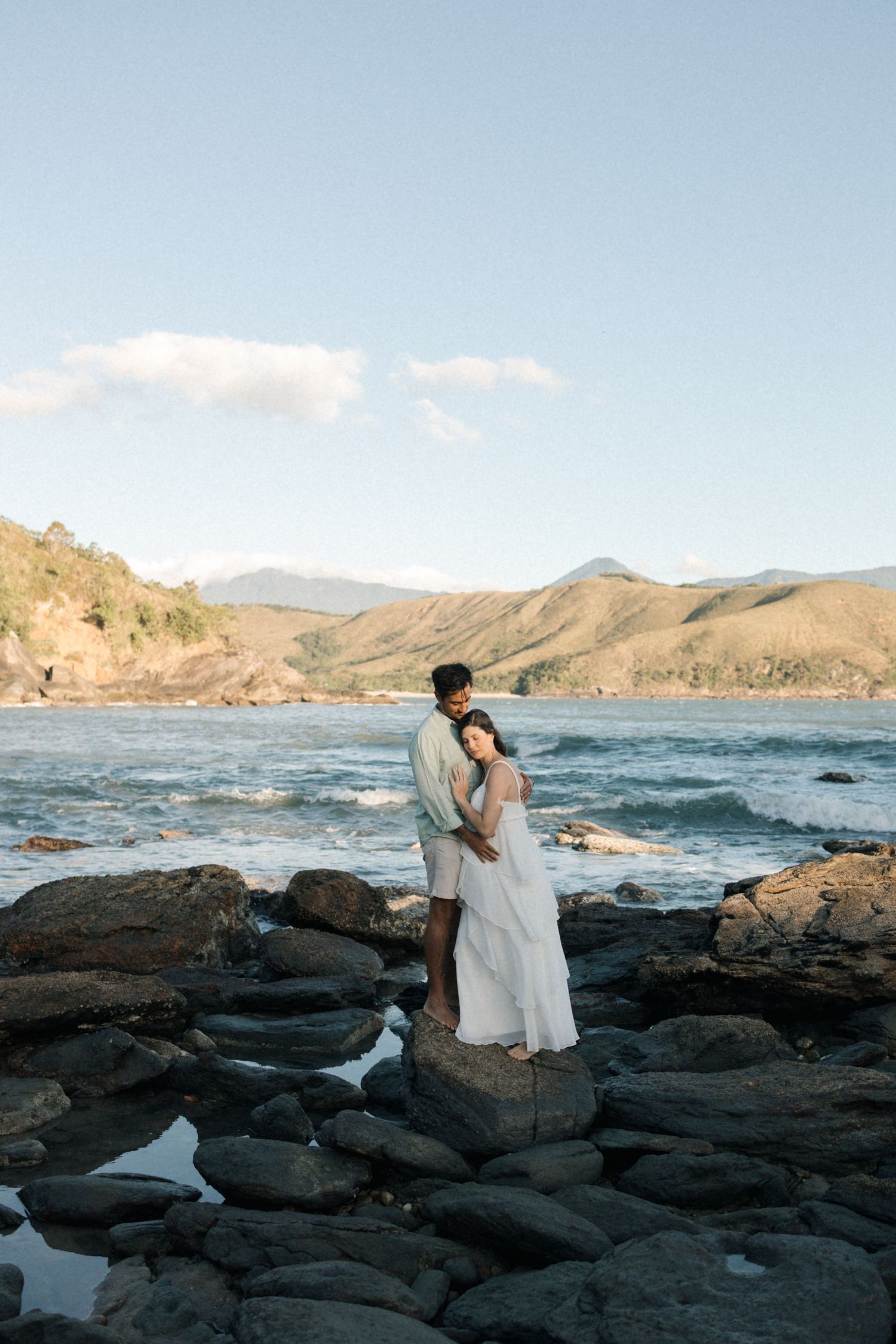 A romantic couple stands on black volcanic rocks by the ocean with mountains in the distance.