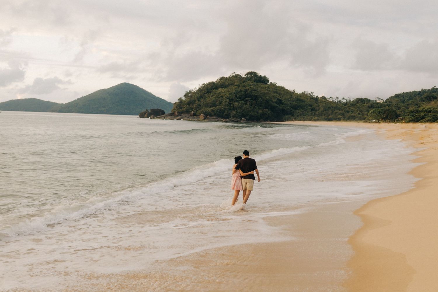 Um casal caminhando por uma praia isolada com montanhas e vegetação exuberante ao fundo em um dia nublado.