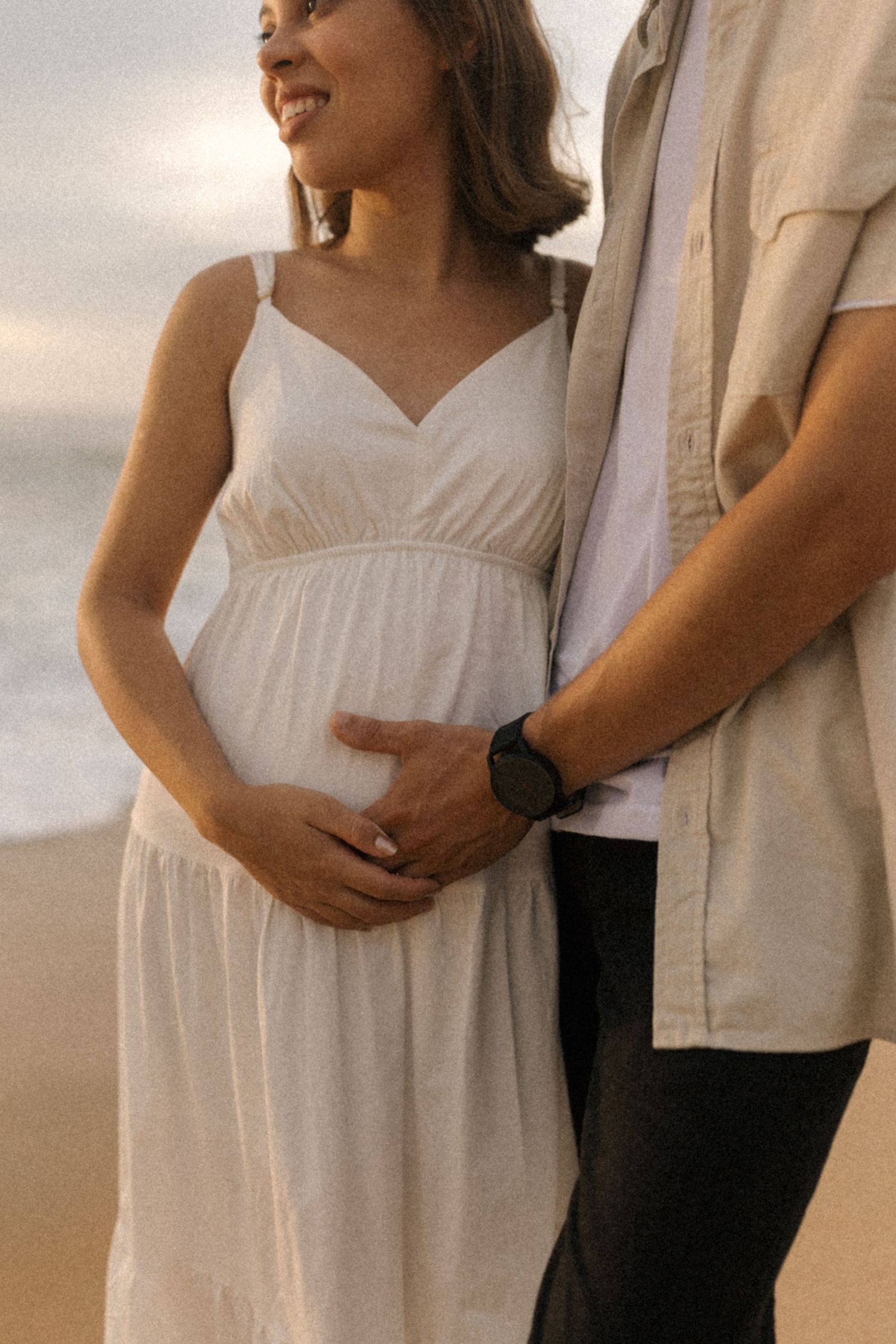 Tender maternity moments captured on a beach at sunset with couple in white and beige outfits.
