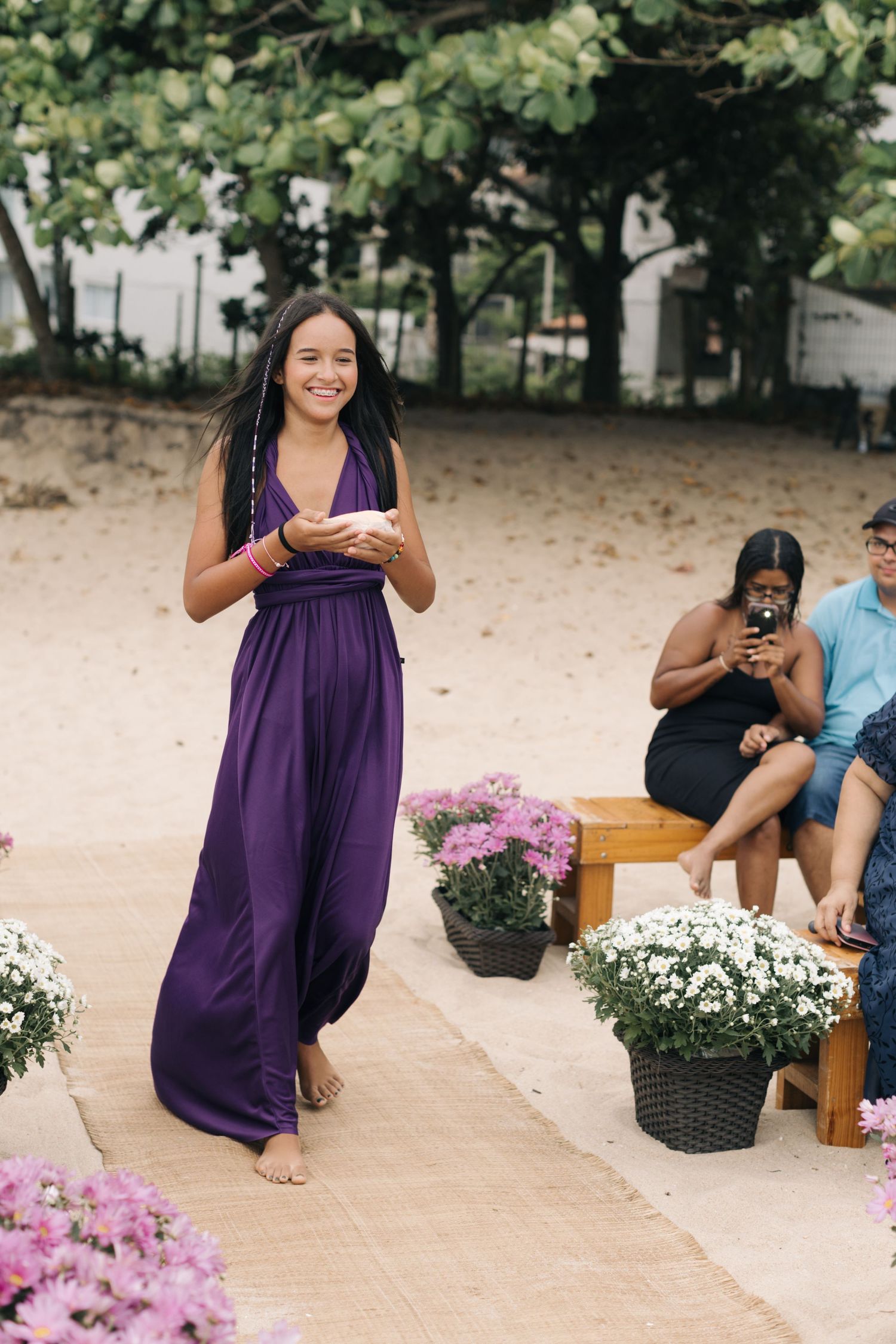 Person in a purple dress kneeling beside pink flowers during an outdoor celebration.