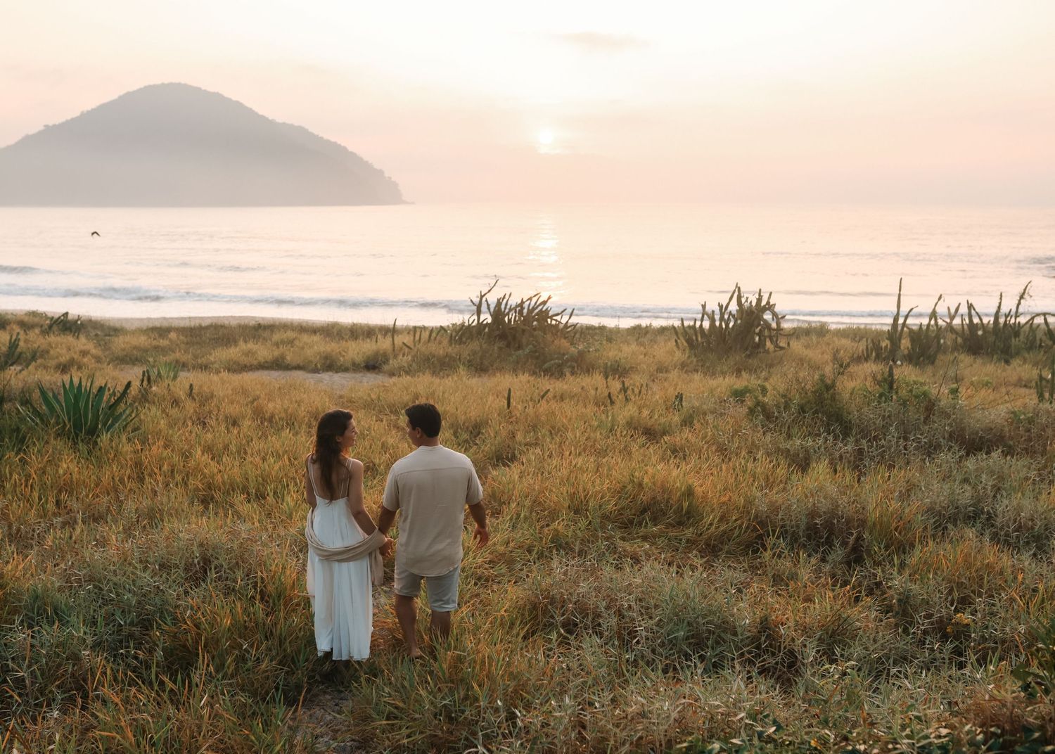 A couple walks hand in hand through a golden field at sunset with mountains in the distance.