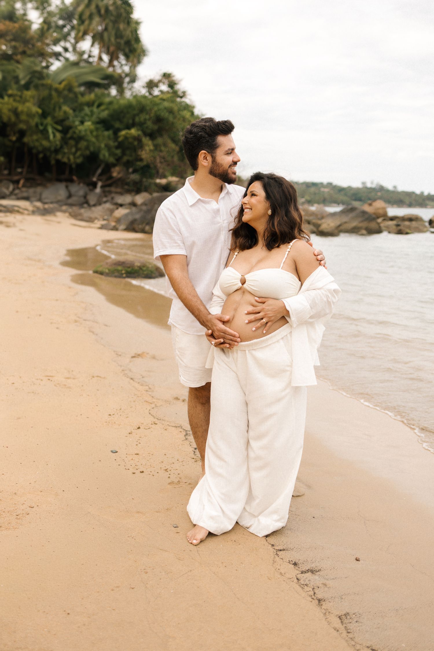 A couple in white outfits shares an intimate moment on a tropical beach during a maternity photoshoot.