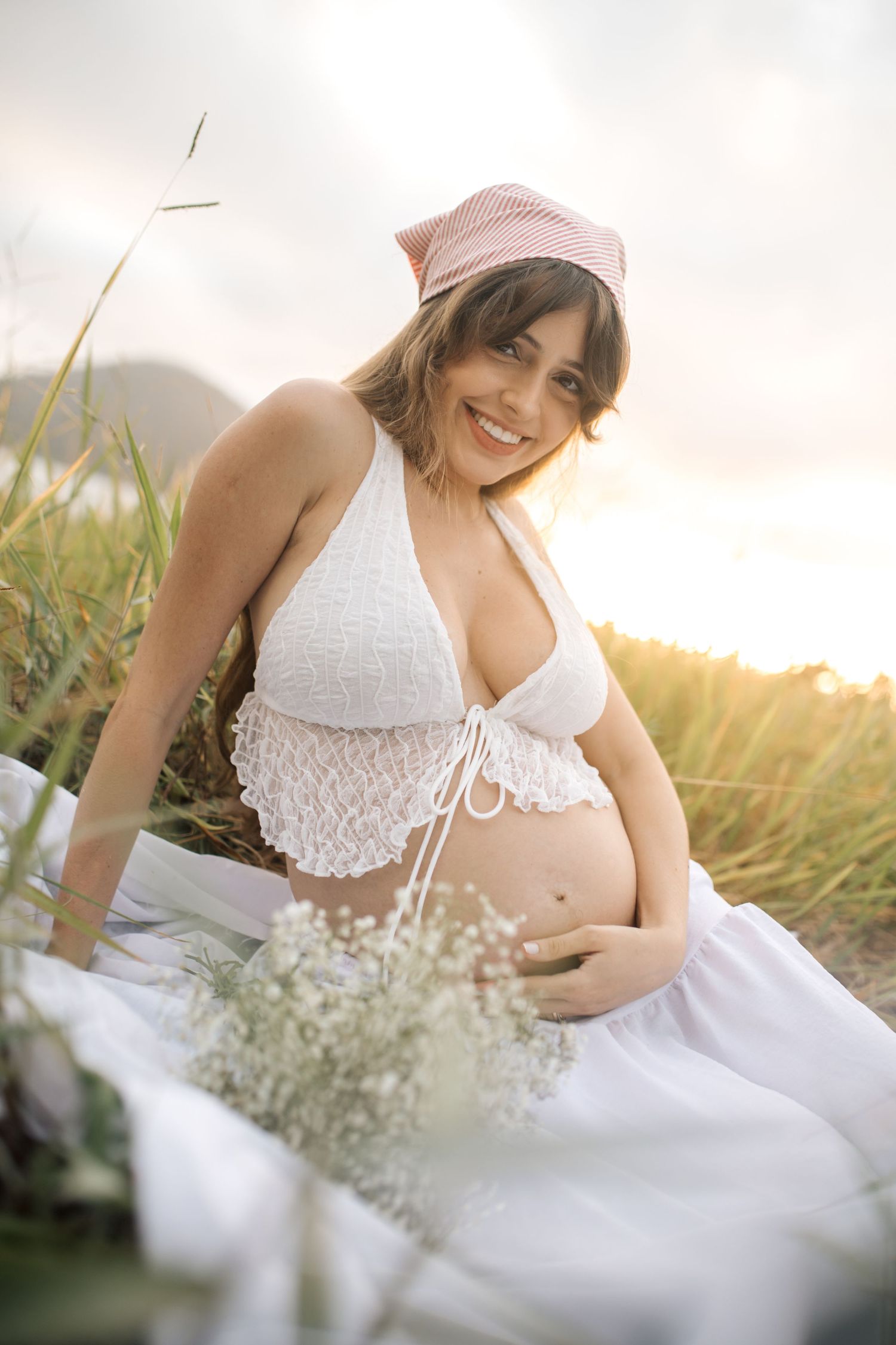 Maternity portrait in a white lace top sitting among tall grass at sunset with delicate flowers nearby.