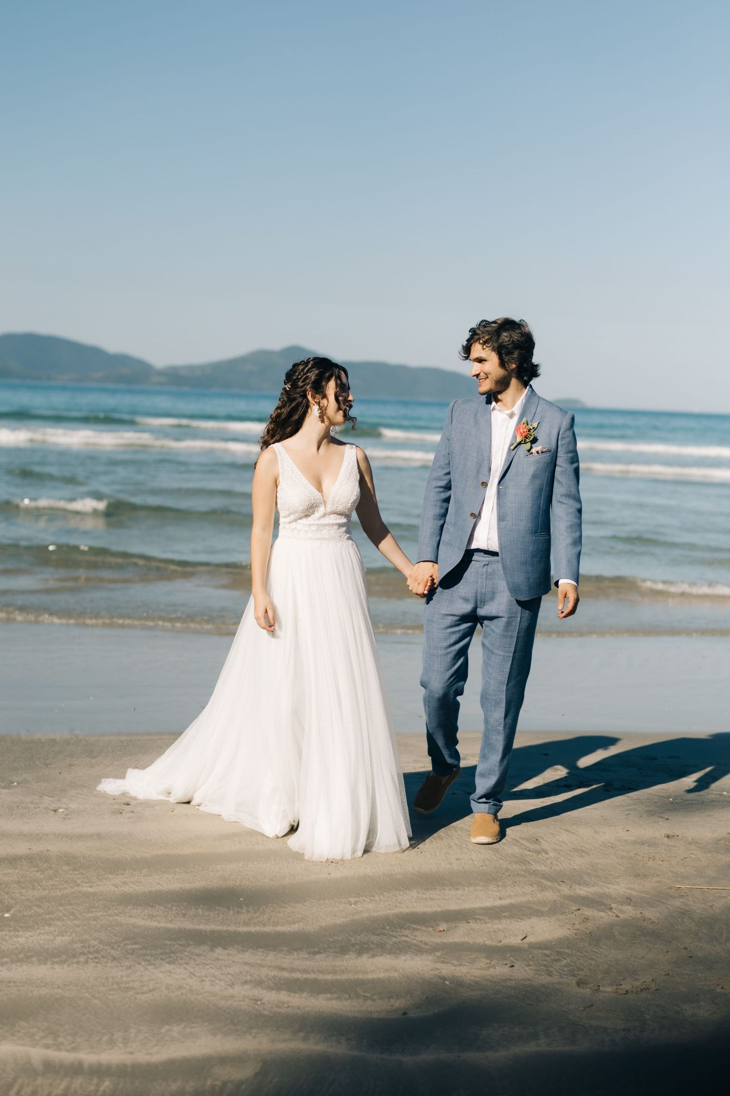 A couple walking on a beach at sunset, with waves and mountains in the background.