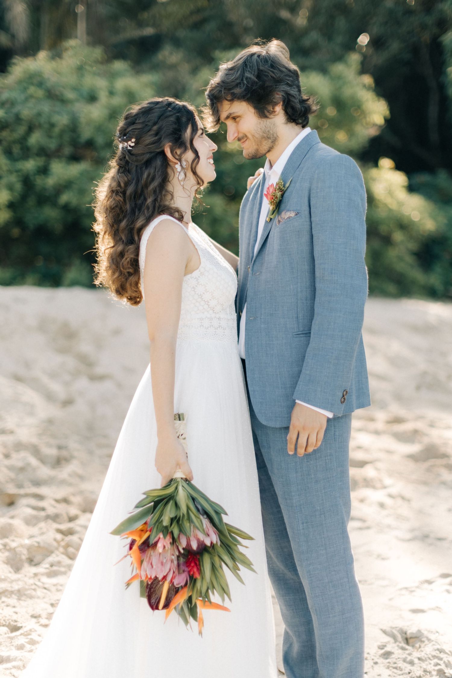 Couple shares romantic moment on beach with tropical bouquet and light blue suit.