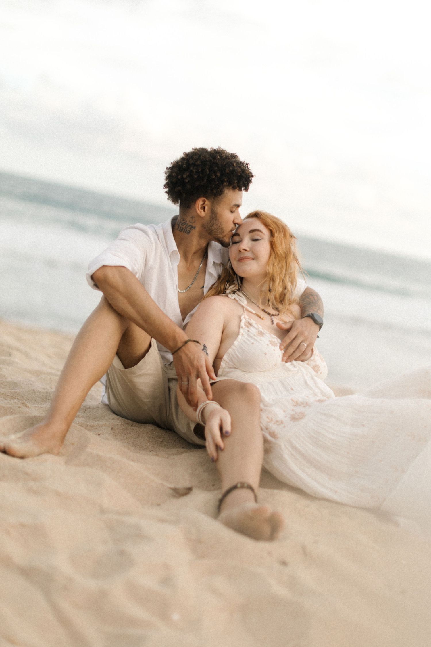 Two people share an intimate moment while lying on the beach sand with soft natural lighting.
