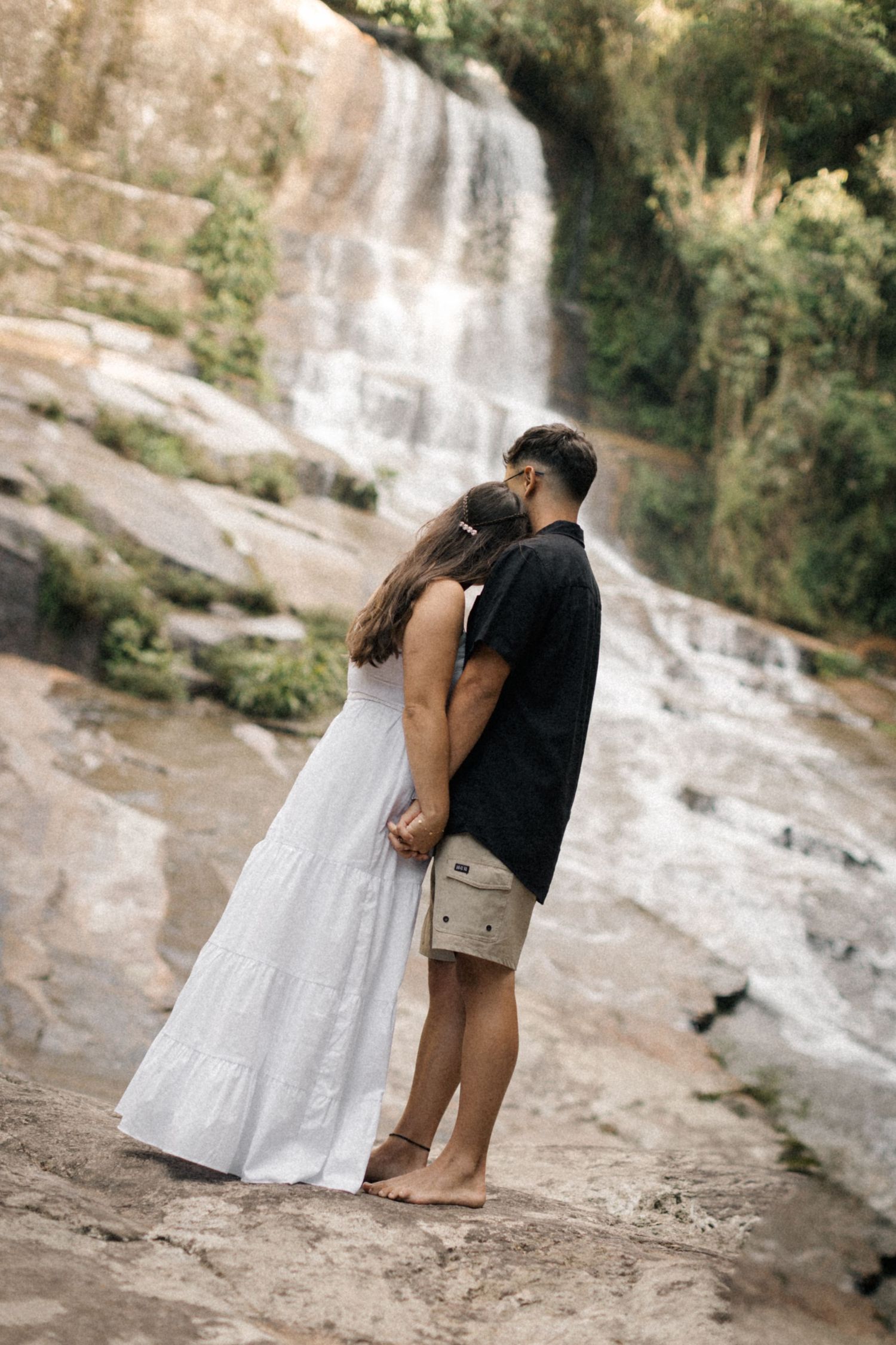 A couple in white and black attire shares romantic moments by a scenic waterfall.