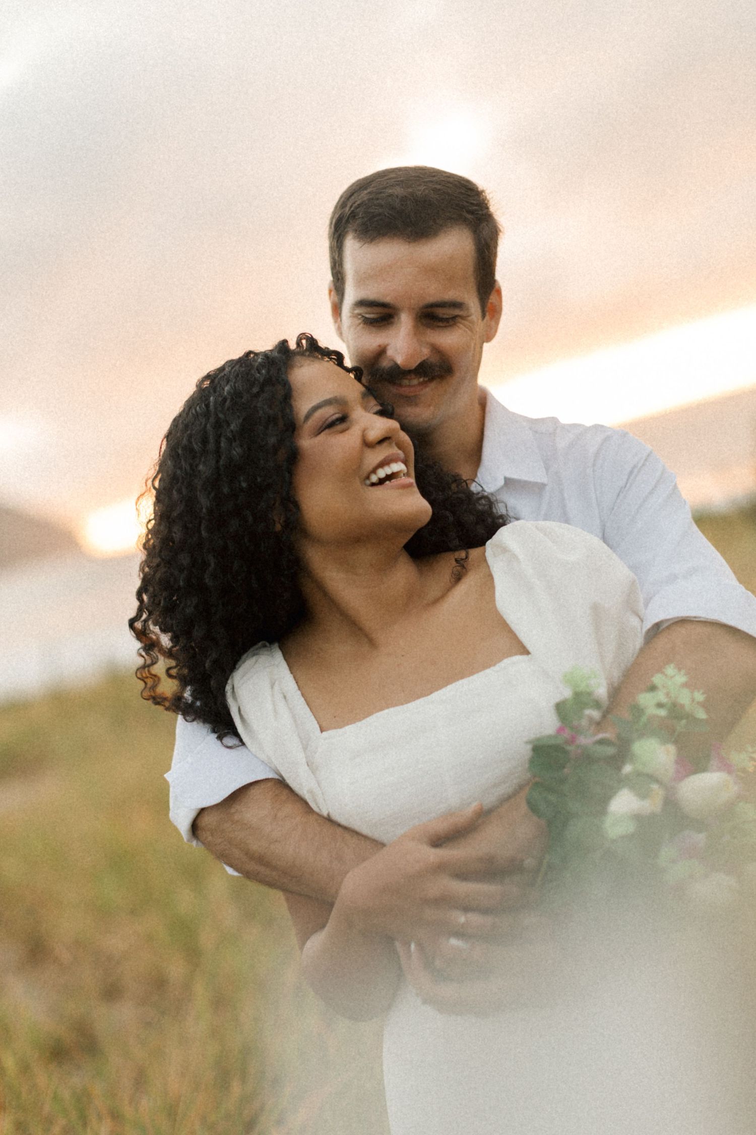 A romantic couple embraces at sunset in a field, with the woman wearing white and laughing joyfully.