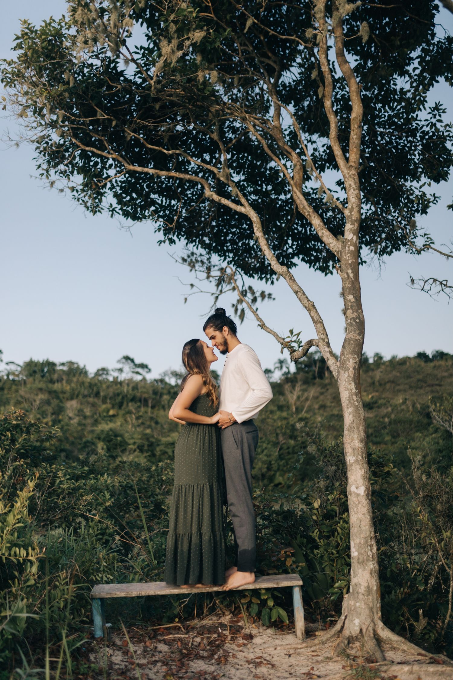 A romantic couple stands on a wooden bench under a large tree in a natural outdoor setting.