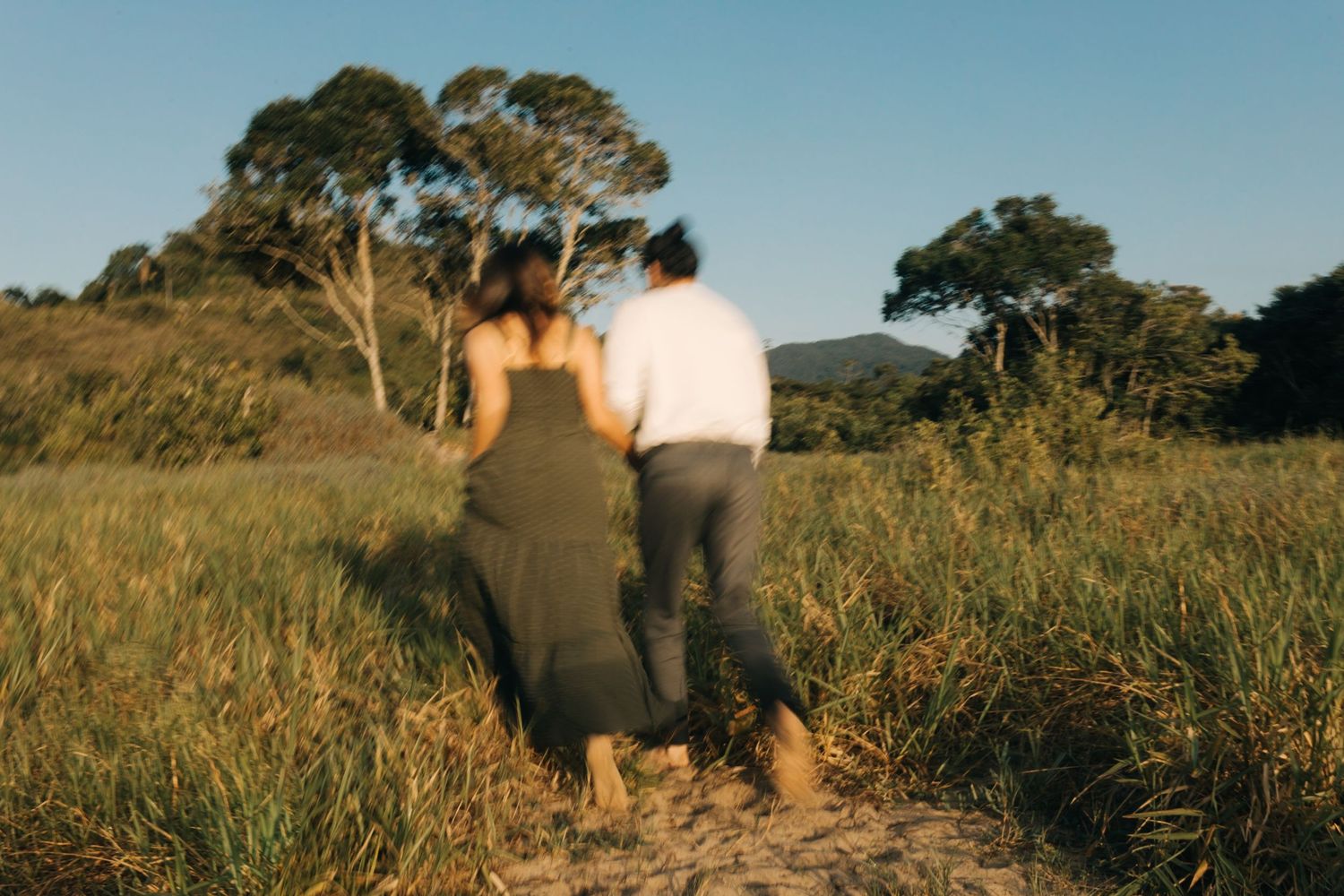 A couple walks away from camera through tall grass at sunset.