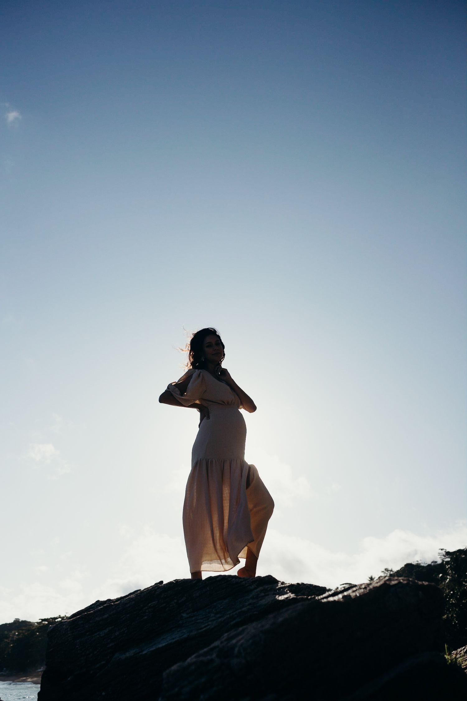 Dramatic silhouette of person in flowing dress posing on rocky cliff against blue sky.