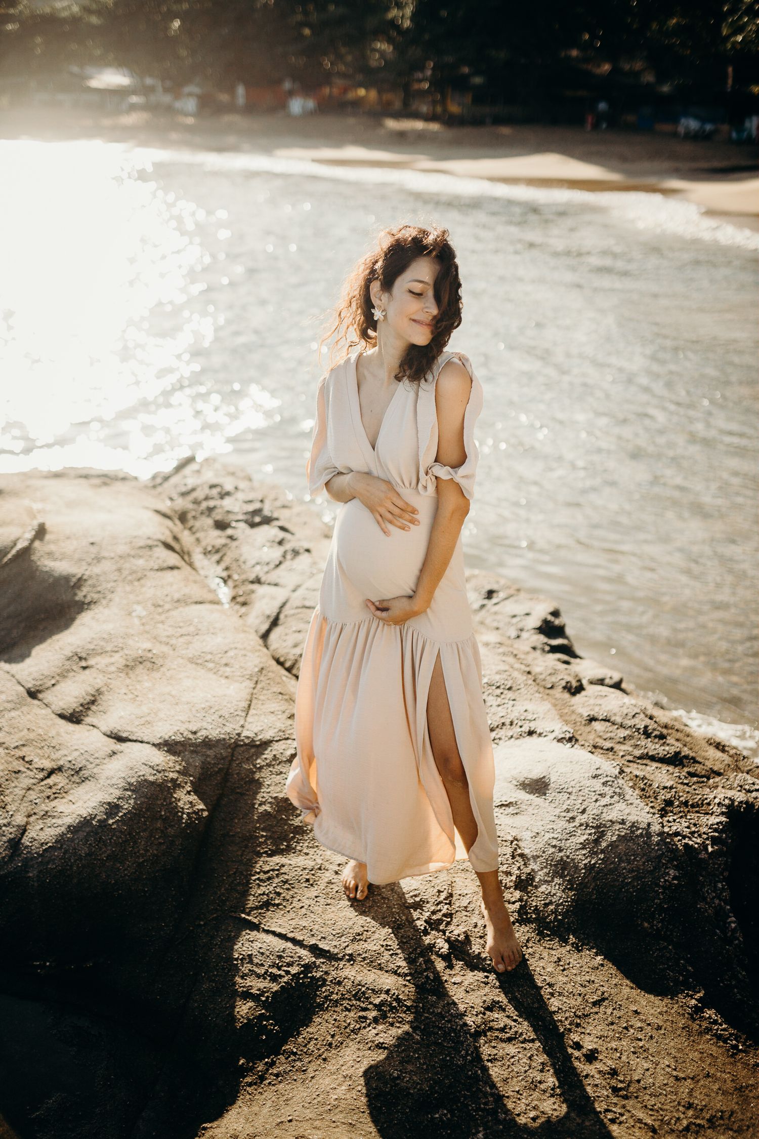 Ethereal maternity photoshoot on a coastal beach with soft sunlight filtering through the rocks.