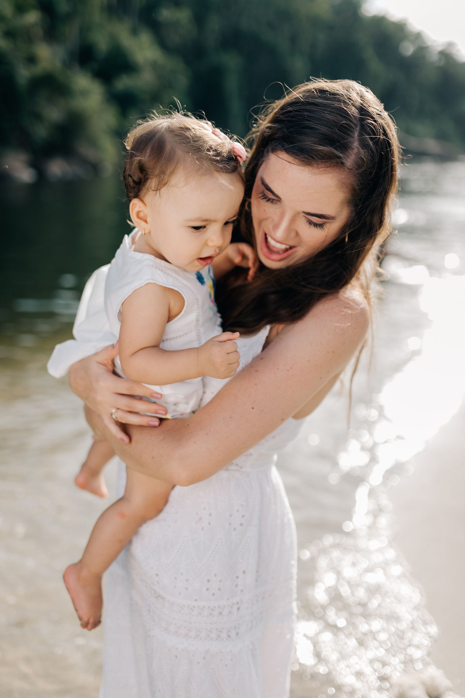A tender moment between parent and child in white dresses by a sunlit lake.
