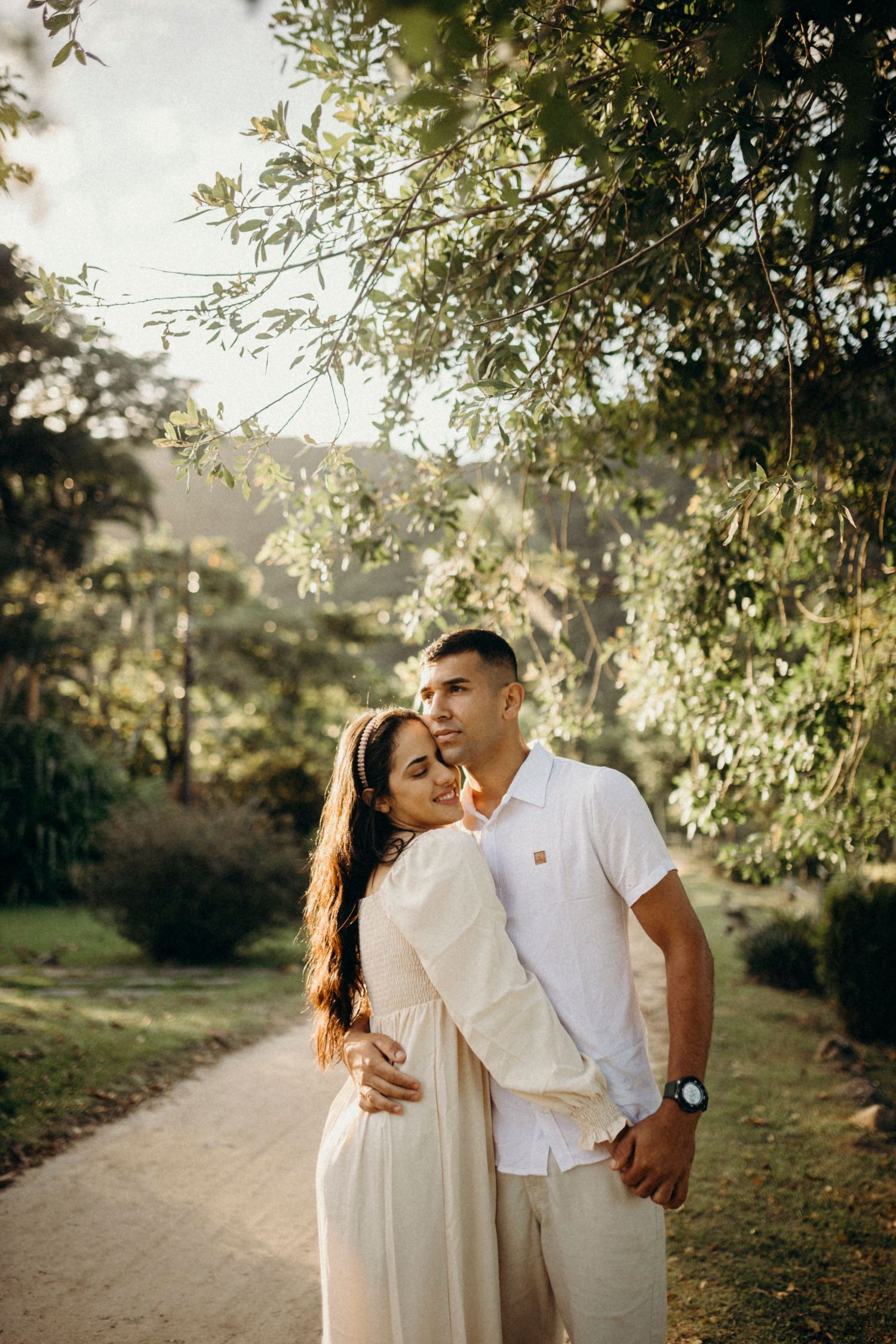 A couple in coordinated white outfits embraces on a sunlit nature path.