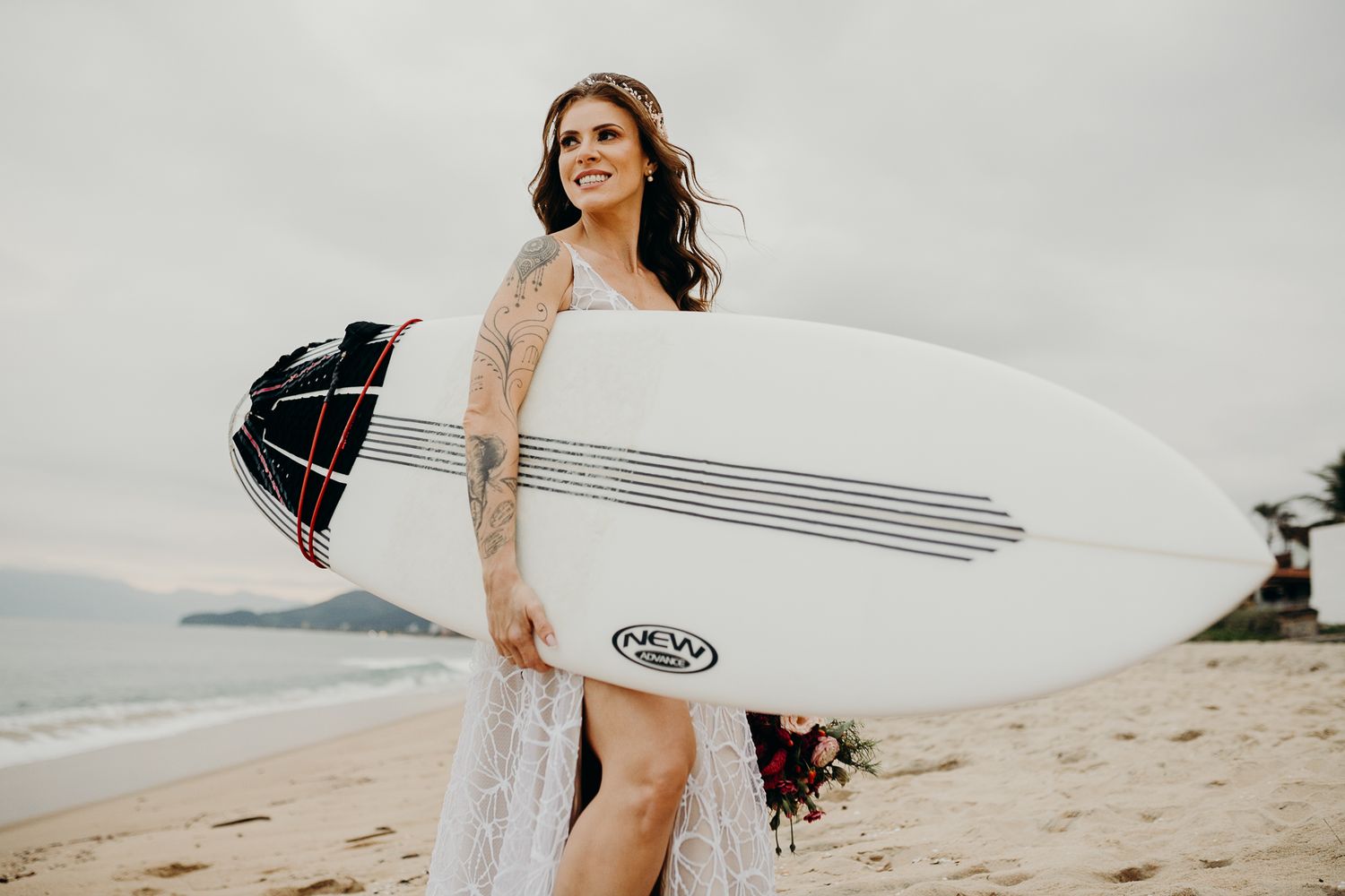 Bride in lace wedding dress holding surfboard on a cloudy beach.