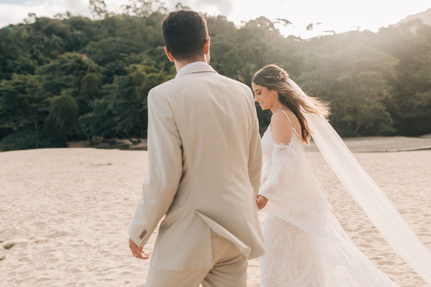 Couple in light-colored wedding attire walking along a sunlit beach with forest backdrop.