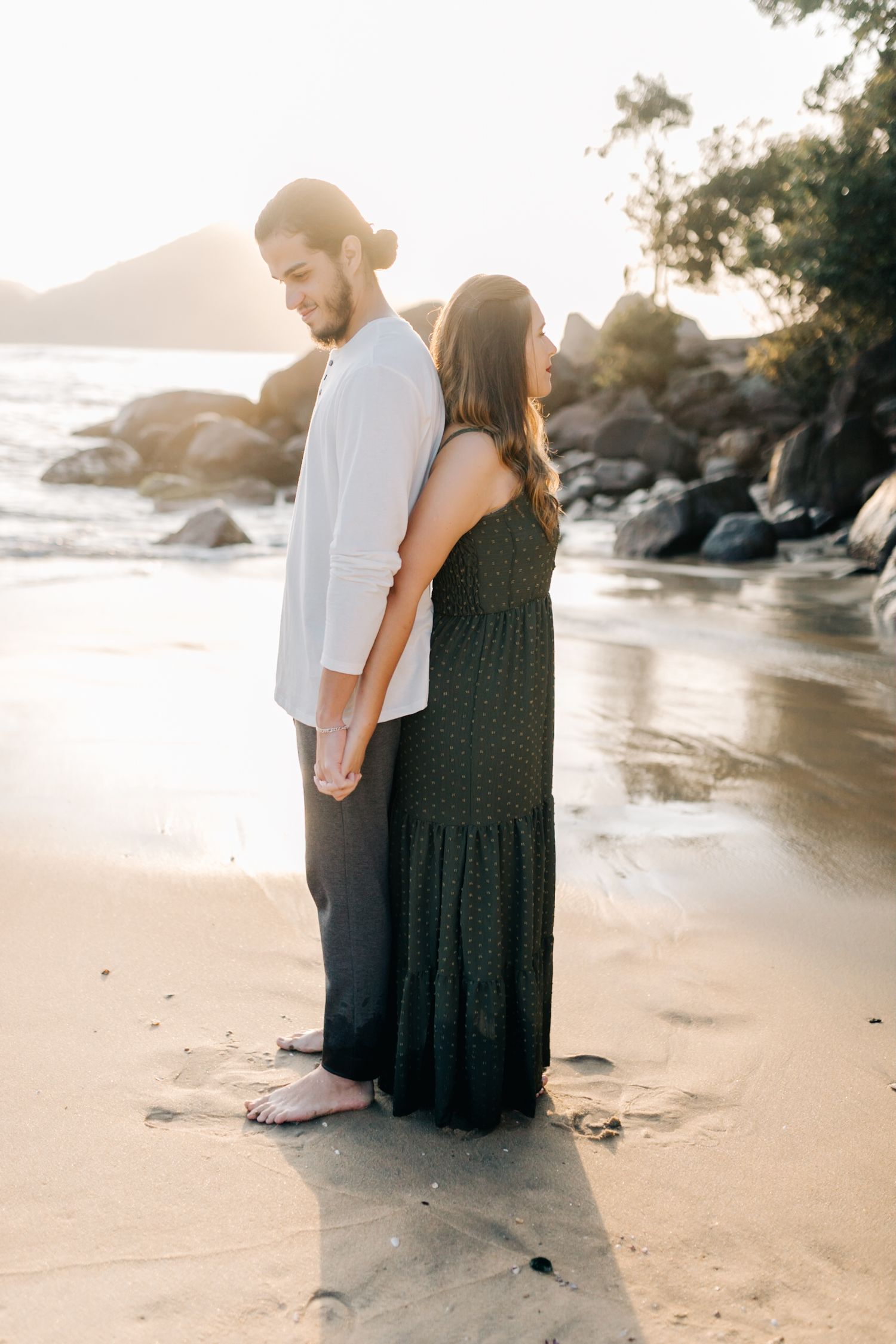 Side-by-side portrait of couple standing on beach during golden hour with rocky coastline behind.