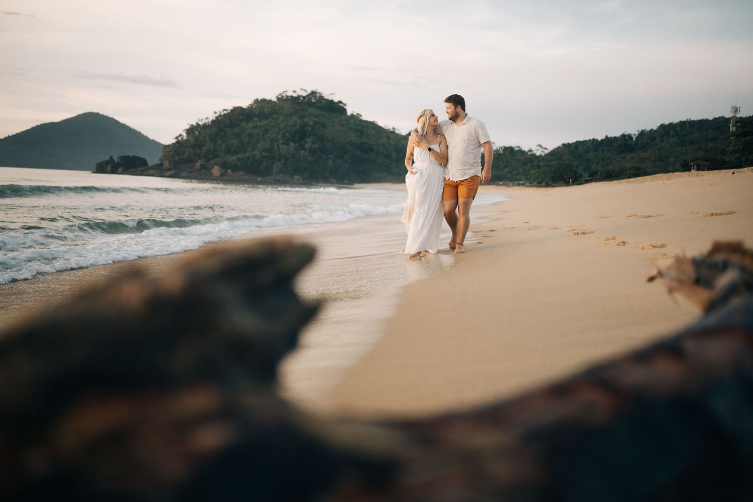 A couple walking along an empty beach with mountains and driftwood in the foreground.