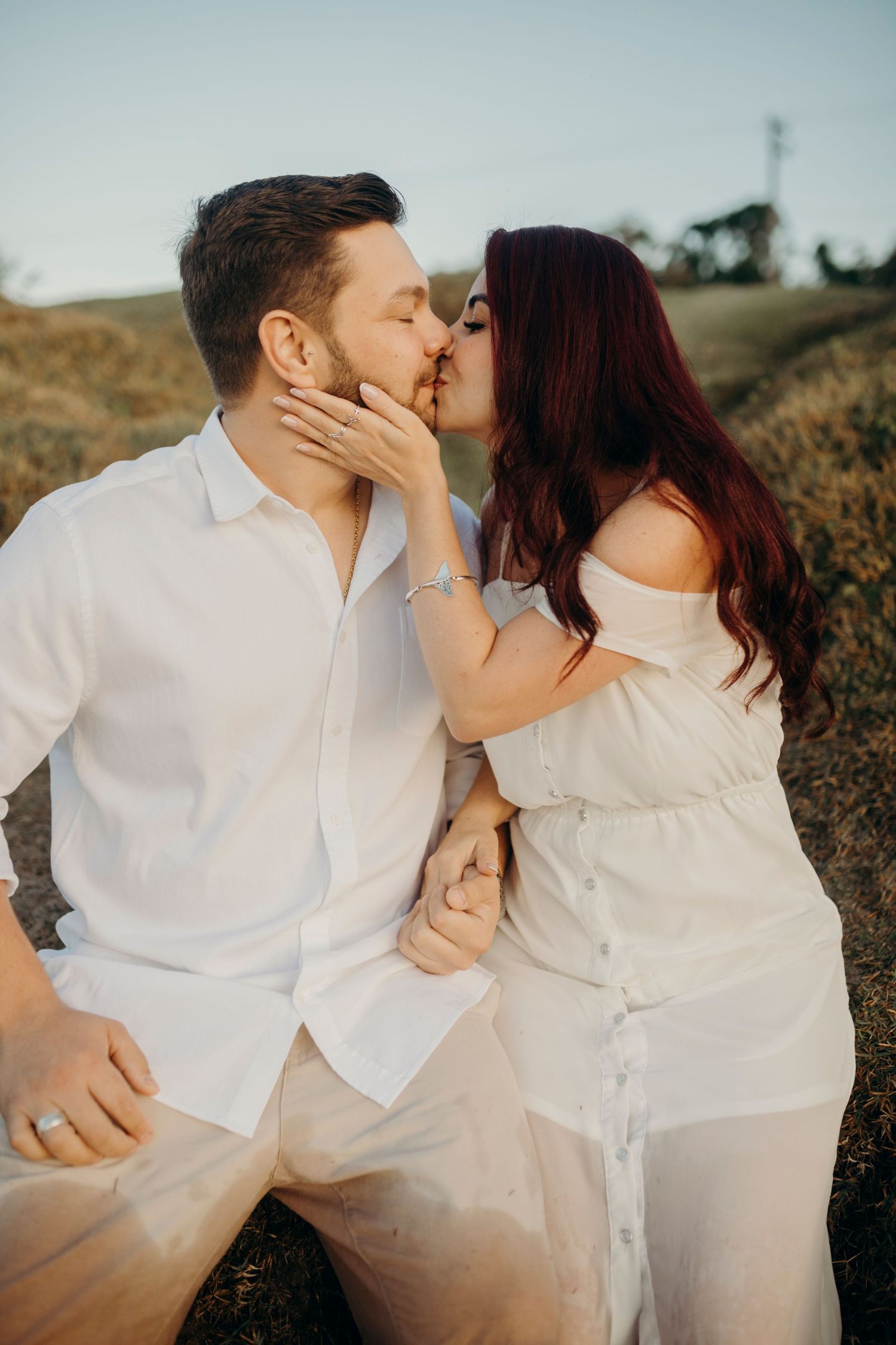 Two people share a romantic kiss while dressed in white against a natural outdoor setting.