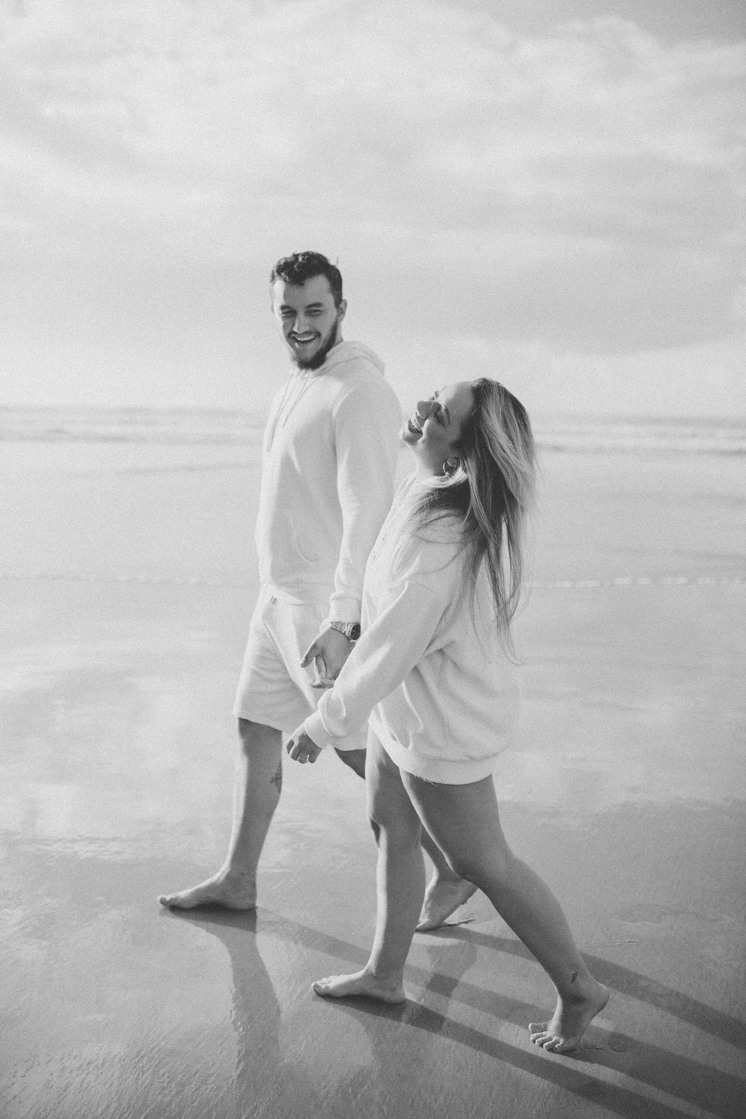Couple in matching white outfits holding hands while walking barefoot on beach.