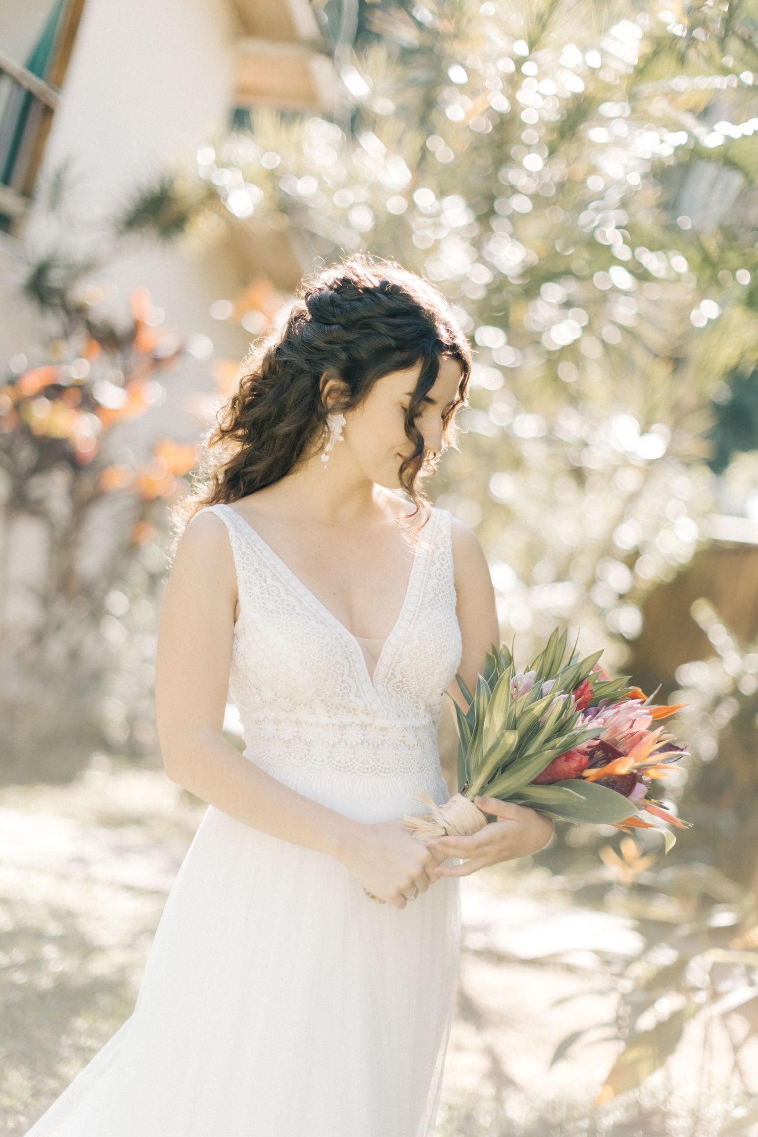Elegant bride in a white lace v-neck dress holds a tropical bouquet in a sunlit garden setting.