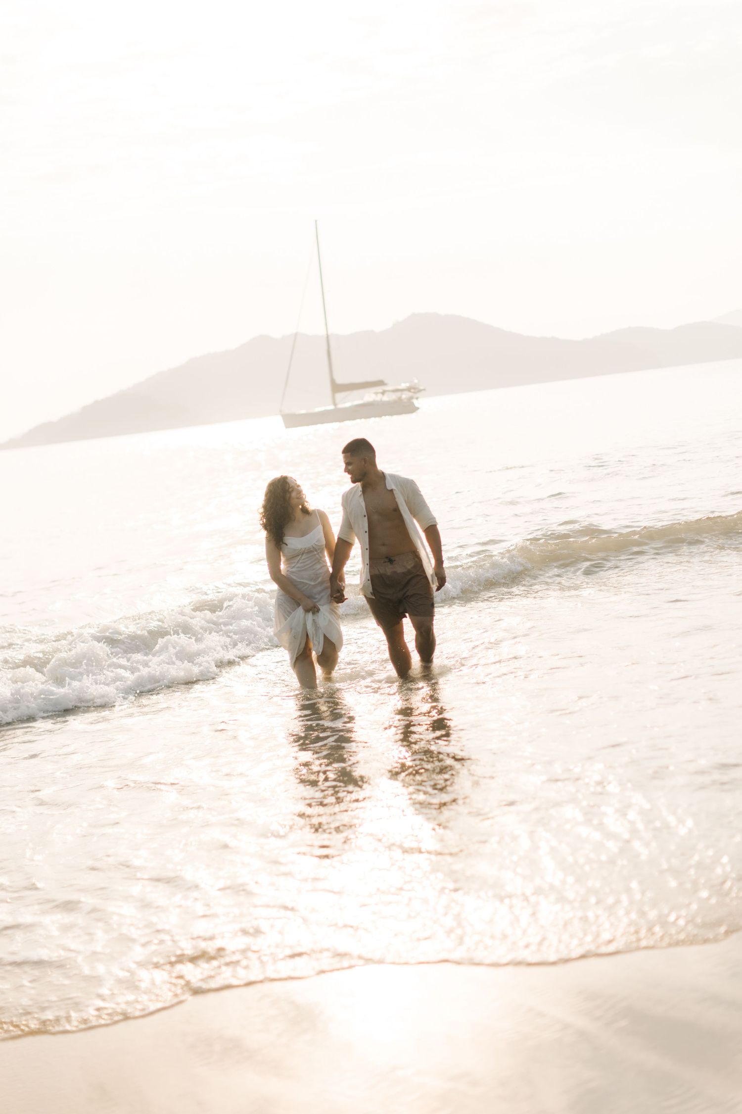 A couple strolling through gentle waves on a beach at sunset.