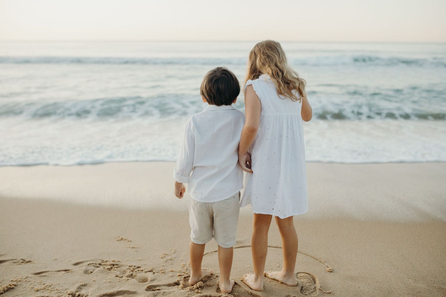 Two children in white outfits playing together on a peaceful beach at dusk.