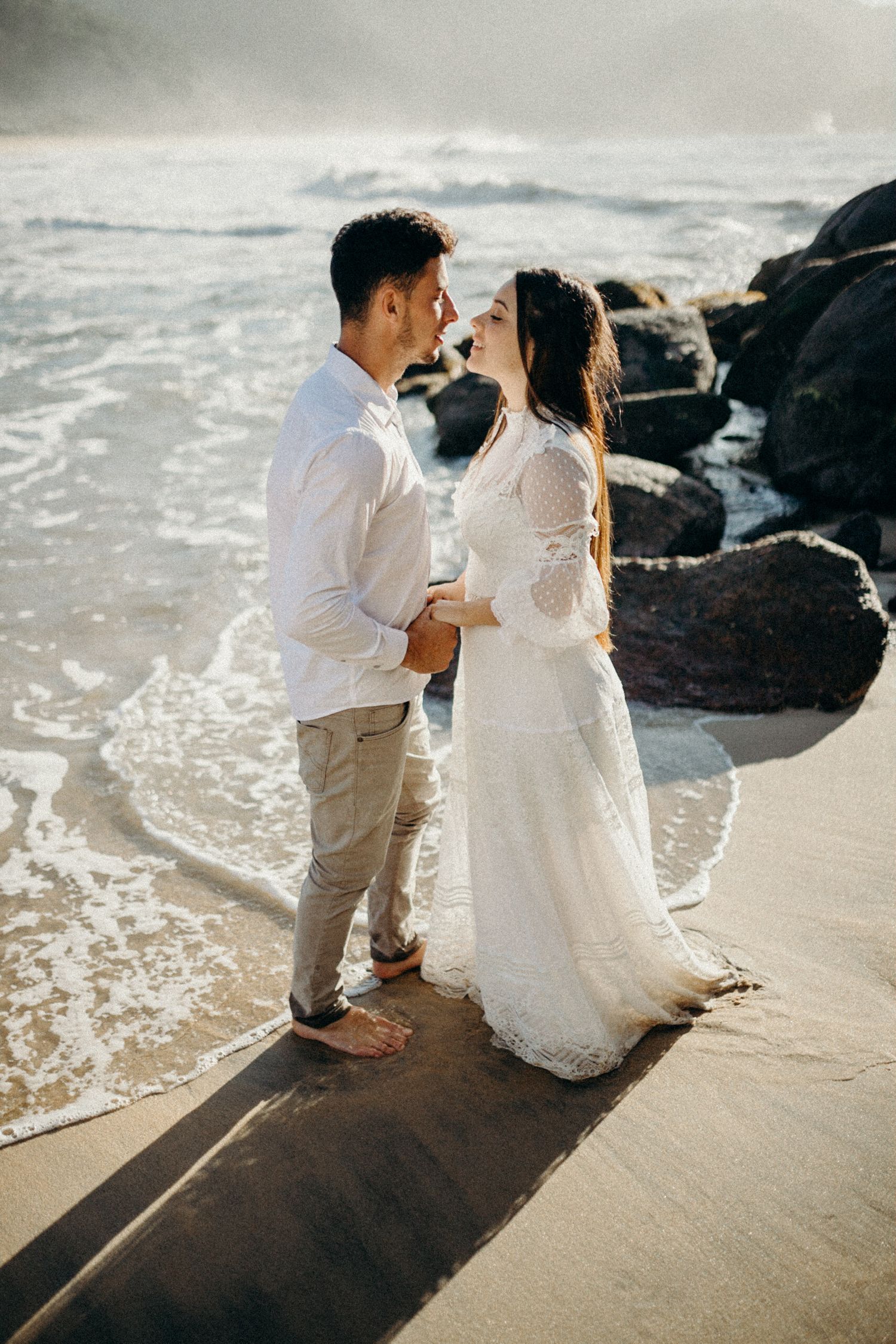 A couple in white clothing stands on rocky beach shore with waves crashing behind them.