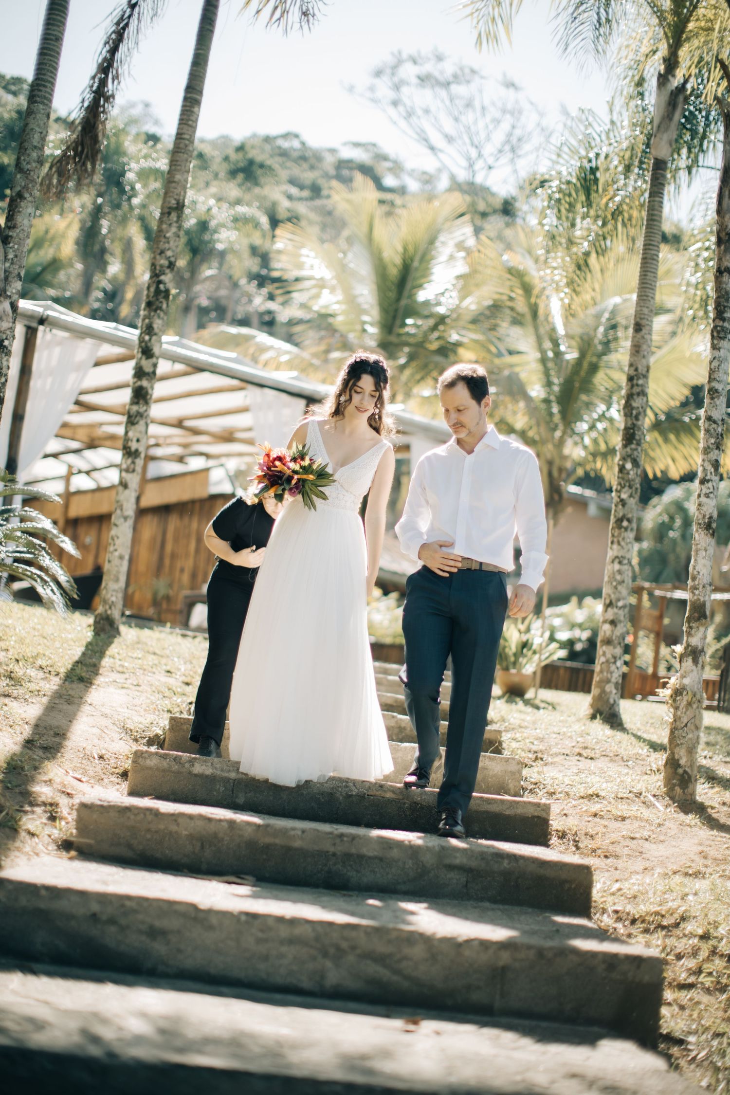 A white bridal gown flows in the sunlight among palm trees at an outdoor wedding venue.