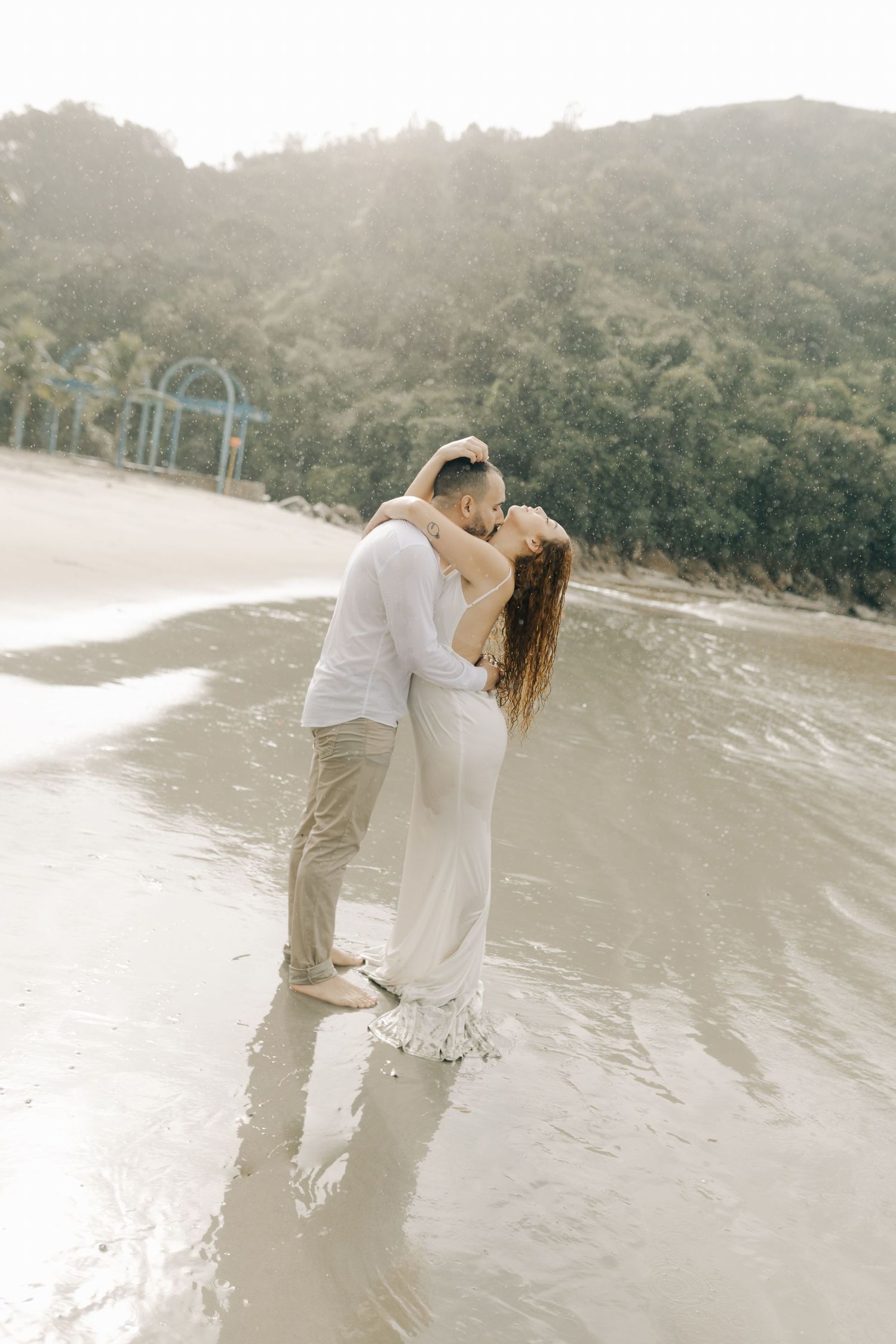 A couple embraces on a scenic beach with lush mountains in the background.