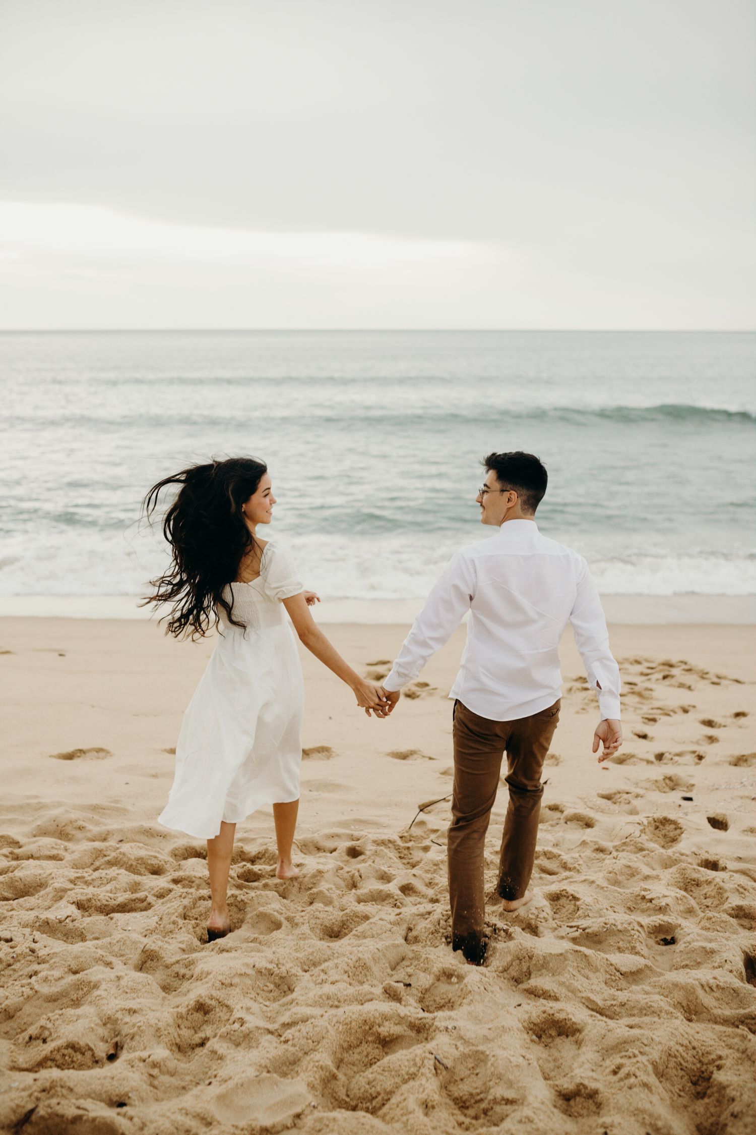 A couple walks hand in hand on a sandy beach with waves crashing behind them.