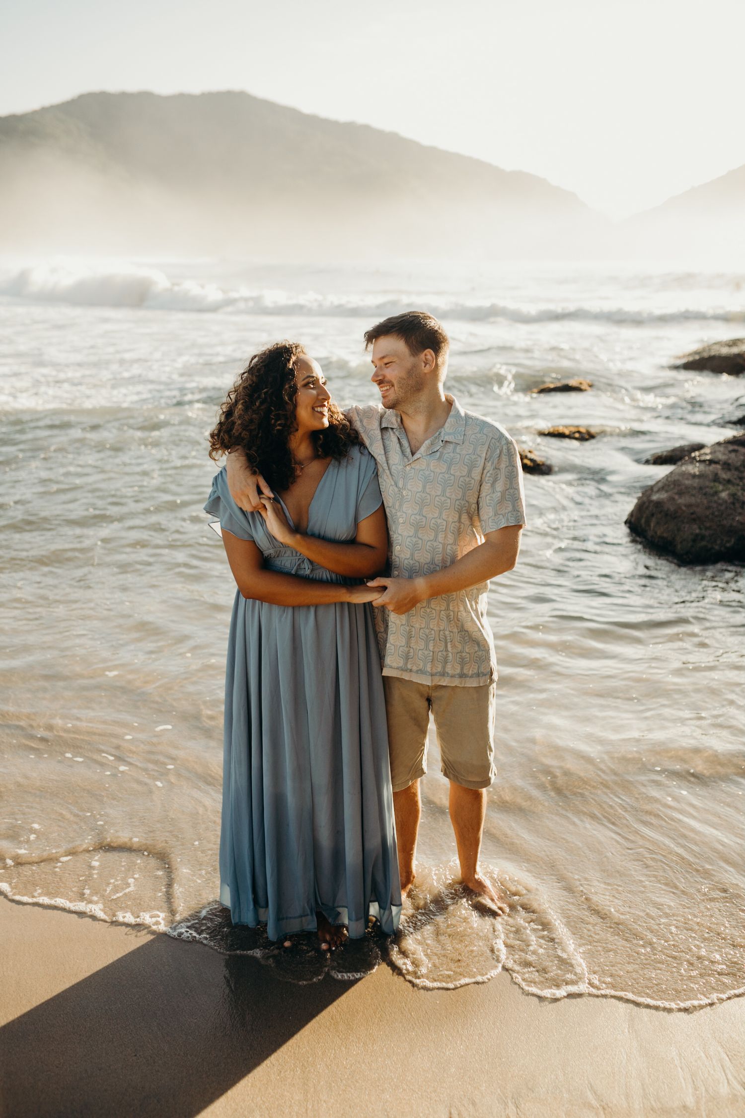Couple walking together on a scenic beach with waves crashing and mountains in the background.