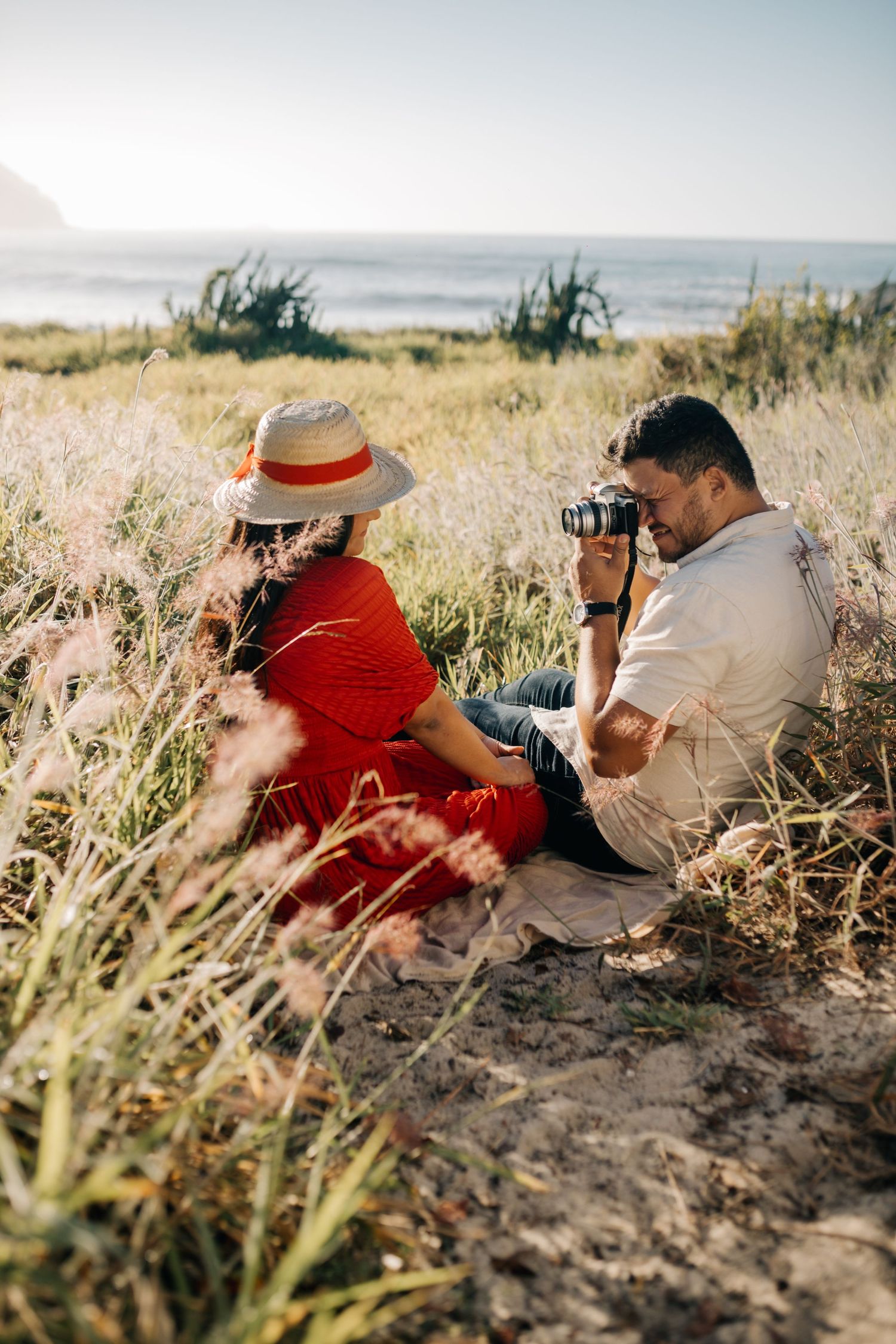 Person in red dress and hat poses while companion takes photos by ocean.